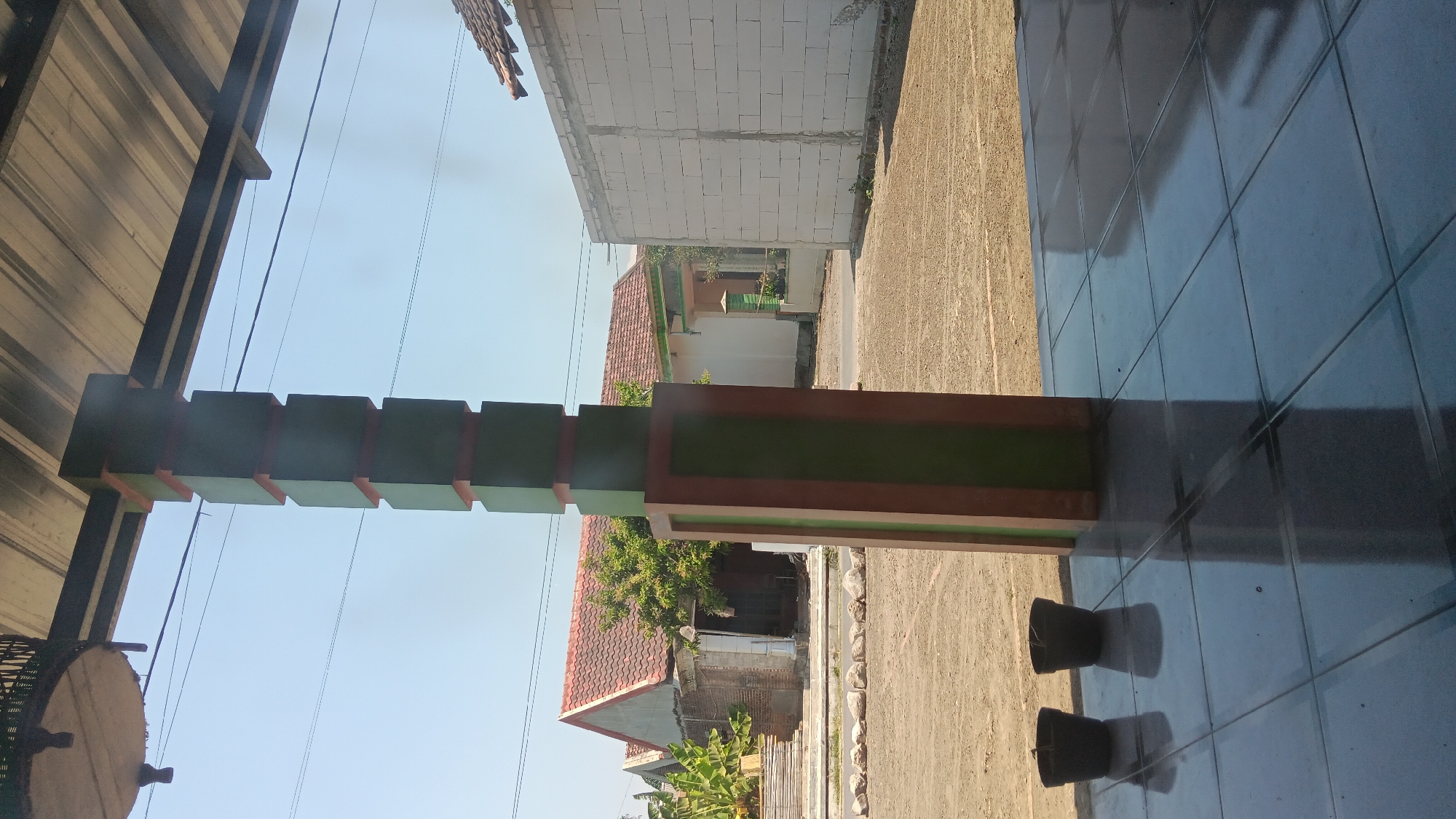 In Sonobekel, Indonesia, a tiled porch area faces a dirt lot with buildings in the background. A prominent pillar, painted in alternating sections of dark green and reddish-brown, stands in the foreground. Two dark, simple pots sit on the tiled floor to the left of the pillar. Beyond the dirt lot, a house with a red-tiled roof and a white structure made of cinder blocks are visible. The sky is clear and blue.