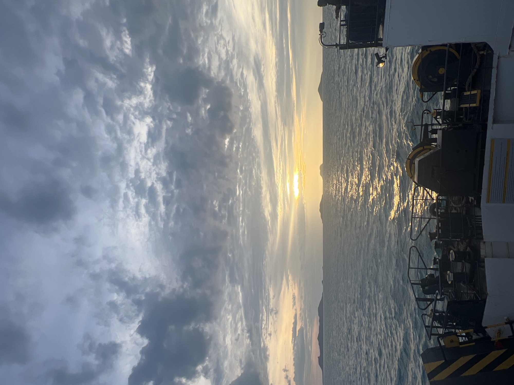 The image shows a view from a ferry at sunset. The sky is filled with dramatic clouds, ranging from dark gray to light gray and hints of yellow from the setting sun. The sun is low on the horizon, casting a golden reflection on the rippling water. Several islands are visible in the distance, silhouetted against the bright sky. In the foreground, the deck of the ferry is visible, with equipment like winches and railings. A person is standing on the right side of the ferry, looking out at the sea. The overall atmosphere is peaceful and serene.