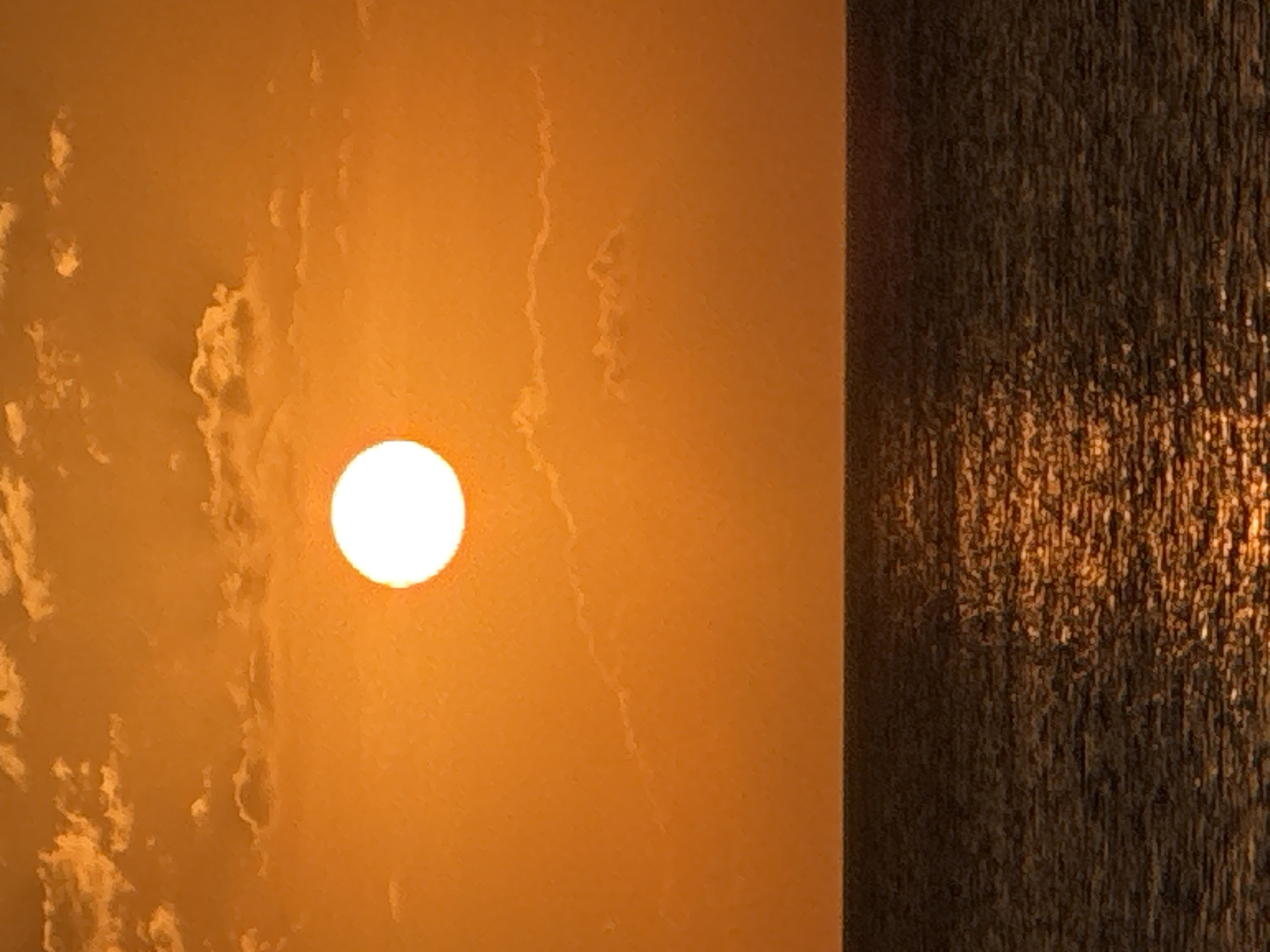 The image shows a sunset over the ocean. The sun is a bright, white orb positioned in the upper center of the frame, casting an orange glow across the sky. Wispy clouds are scattered throughout the sky, tinted orange by the setting sun. The ocean occupies the lower portion of the image, appearing dark and textured with the reflection of the sun creating a shimmering path of light across its surface. The horizon line is clearly defined, separating the sky from the sea. This scene is typical of sunsets witnessed in Kuta Selatan, Indonesia, a region known for its beautiful beaches and ocean views.