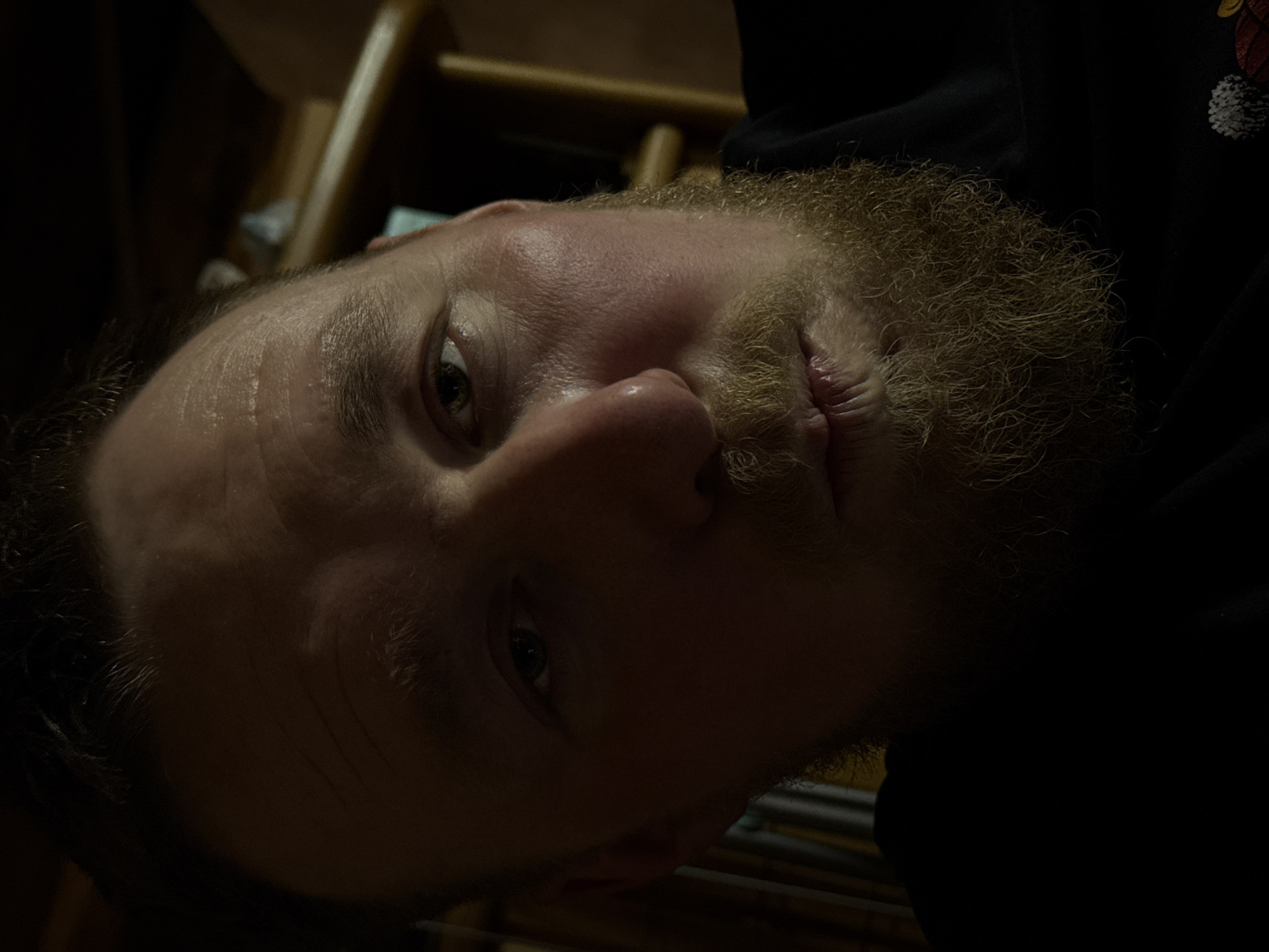 A close-up, low-angle shot of a bearded man. The man has light skin and a blond beard. The background is dark, with a blurry wooden structure. The location is not determinable.
