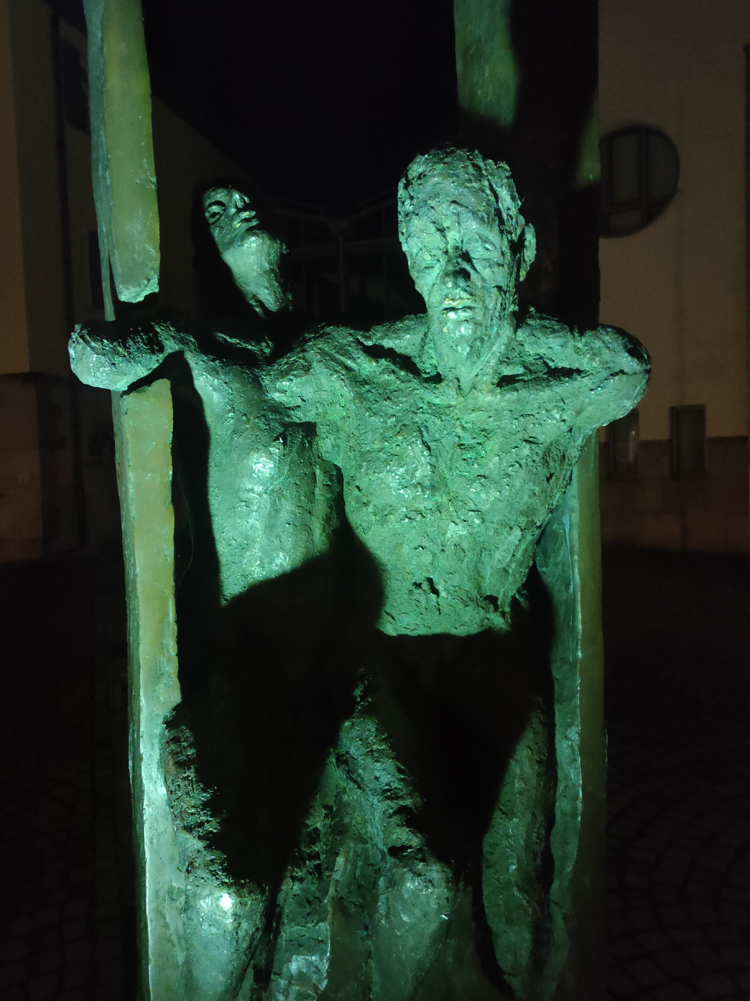 This is a close-up, low-angle, night-time photograph of a weathered, green-patinated bronze sculpture located in Kirchheim am Neckar, Germany. The sculpture depicts two figures, their bodies intertwined and distorted. The figure in the foreground, on the right, is a male torso with a heavily textured and aged surface, his face visible with closed eyes and a grim expression. His arms are extended outwards, appearing to be supported by or pushing against vertical elements of the sculpture. Behind him and slightly to the left, another figure, possibly female, is partially visible. This figure's head is tilted back, her face also showing signs of wear and distress. The lighting appears to be artificial, casting dramatic shadows and highlighting the rough texture of the bronze. The background is dark, with hints of a building and cobblestone pavement visible. The overall impression is one of suffering, struggle, or imprisonment. This sculpture is located at the entrance to the town hall ("Rathaus") in Kirchheim am Neckar.