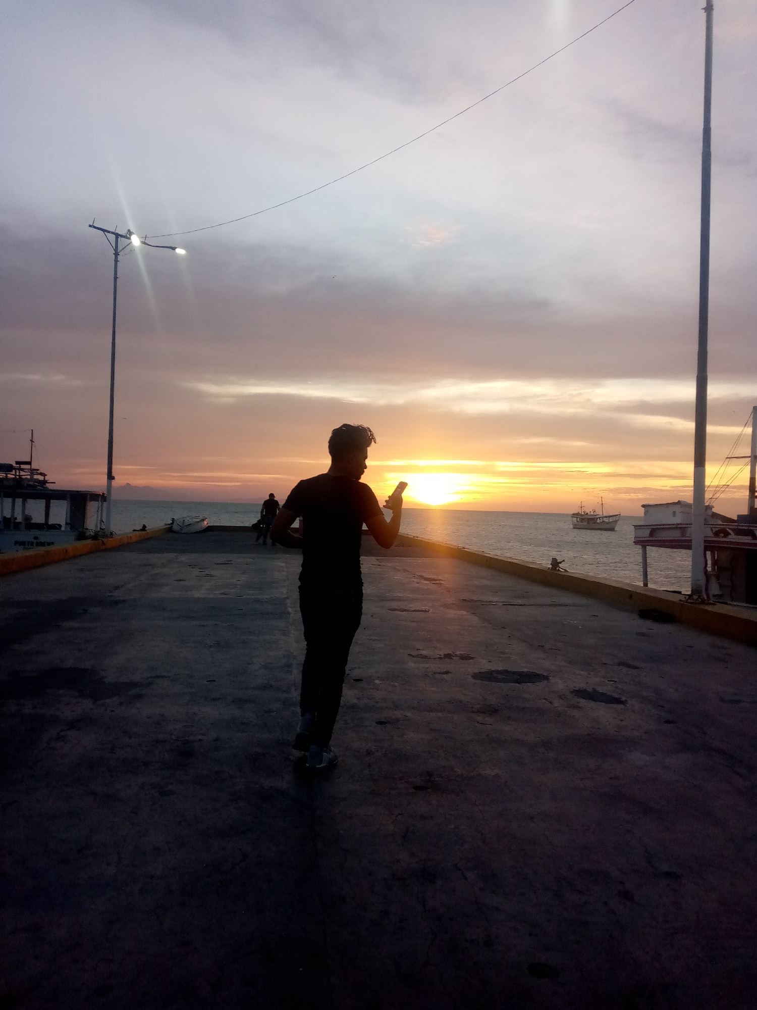 The image depicts a person, facing away from the viewer, standing on a concrete pier or dock, holding a mobile phone. The person is silhouetted against the backdrop of a sunset over the ocean. The pier is lined with boats on either side. In the background, there is another person walking on the pier and a boat at sea. The scene is lit by a street lamp on the left and a tall pole on the right.