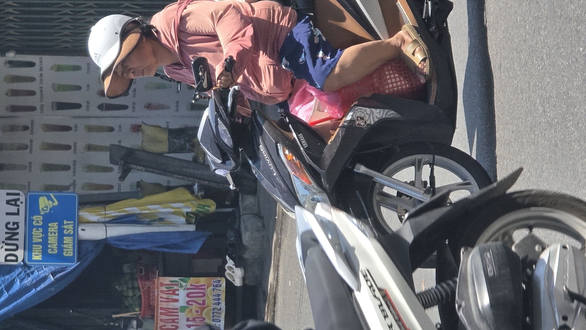The image captures a woman riding a motorcycle in Nha Trang Ward, Vietnam. She is wearing a white cap with a visor and a light pink shirt, and is seen in the rider's seat. She has sandals on her feet, which are placed on the back of the seat. The motorcycle appears to be a Yamaha Luvias. In the background, there's a storefront, likely selling beverages or food, indicated by the sign "CAM VAT 15-20k 0772 444 764." A blue sign indicates "KHU VỰC CÓ CAMERA GIÁM SÁT," which translates to "Area Under Camera Surveillance." Umbrellas are visible near the storefront.