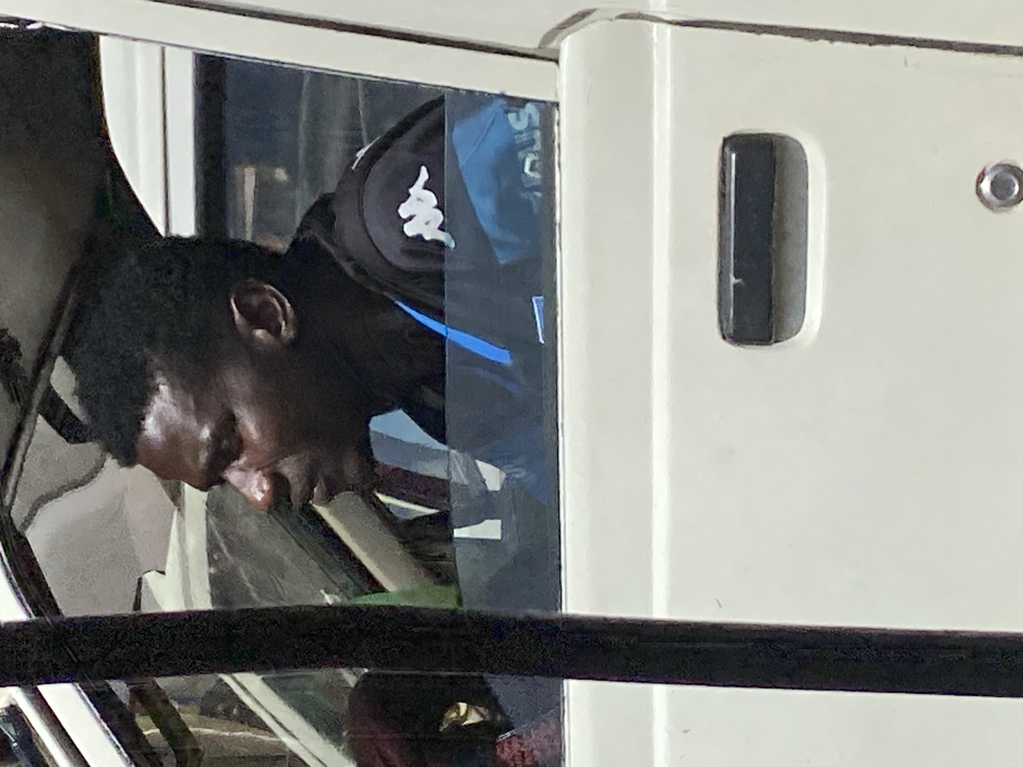 A person is visible, with dark skin and short, dark hair. They are leaning forward, with their head down, and their eyes are not visible. They are wearing a black cap with a white logo, which appears to be the Kappa logo. The background consists of a white surface and a dark, reflective surface, possibly a window or a screen, which shows reflections of the surroundings. The scene seems to be indoors, possibly within a vehicle or a building with large windows. The lighting is somewhat dim, suggesting it might be evening or overcast. There are hints of other objects in the background, including what looks like seats and structural elements. The overall impression is that of someone pausing or resting in a confined space. The location is identified as Bukuru, Nigeria. No specific activity or event is clearly depicted, other than the subject's posture suggesting rest or observation.