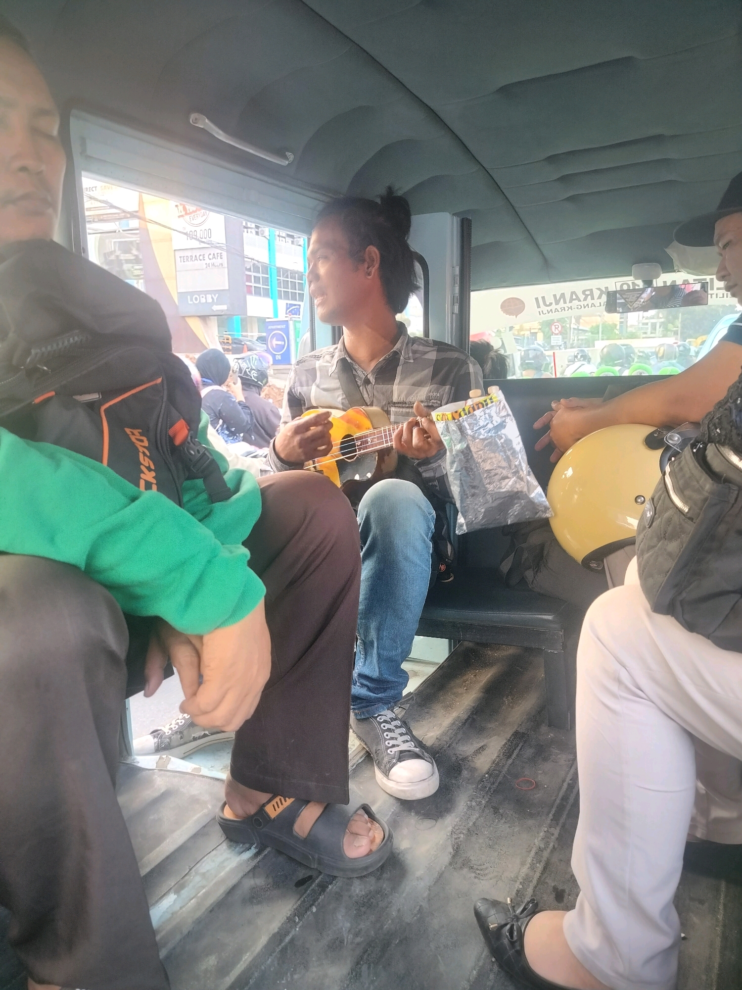 The image shows the interior of a vehicle, possibly a van or jeep, filled with people. In the foreground, a person wearing a green hoodie and black pants is seated on the left. To their right, another person is playing a ukulele. This person is wearing a plaid shirt and blue jeans. To the right of the ukulele player, a yellow helmet and a black bag are visible on a seat, along with the legs of another person wearing white pants. Outside the vehicle, buildings and a street scene are visible through the windows, suggesting an urban environment. One building has signage that appears to be in Indonesian. The overall atmosphere appears casual and communal. The scene is consistent with being in East Jakarta, Indonesia, a densely populated urban area known for its bustling streets and diverse transportation methods.