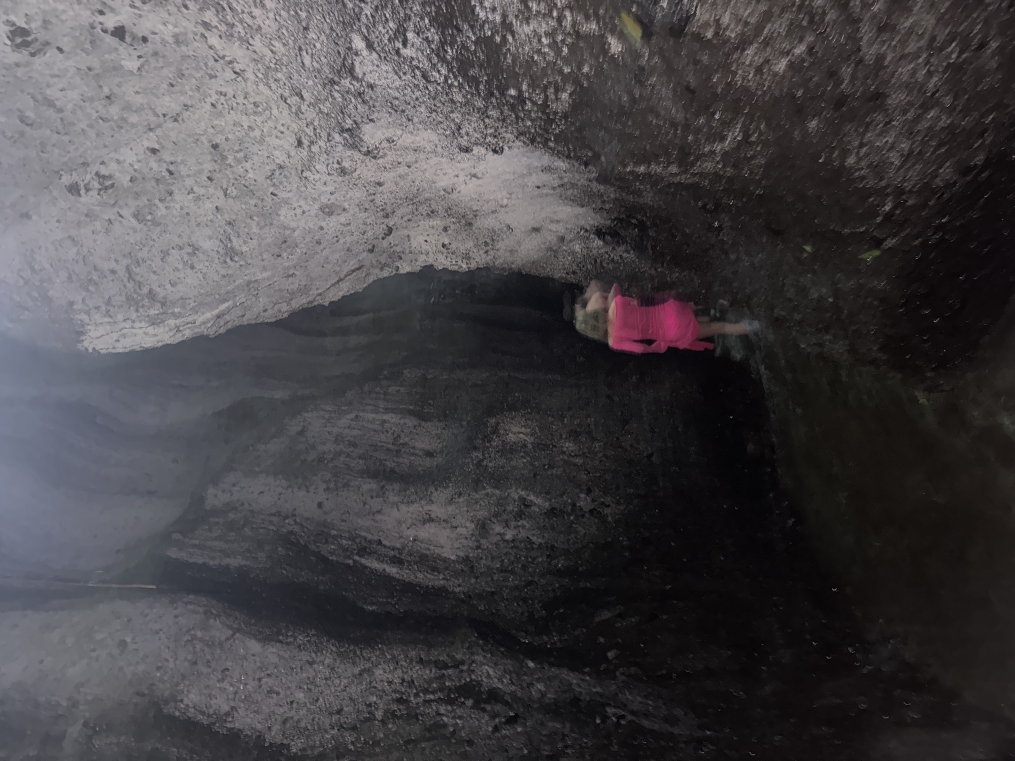 An individual wearing a bright pink dress is walking through a narrow canyon with wet, textured rock walls. The canyon floor appears to be covered in shallow water. The lighting is dim, suggesting an overcast day or the natural low light within the canyon. The scene is located in Bangli, Indonesia, likely at the Tukad Cepung Waterfall or a similar natural formation in the area. The individual is the sole subject, moving away from the camera deeper into the canyon.