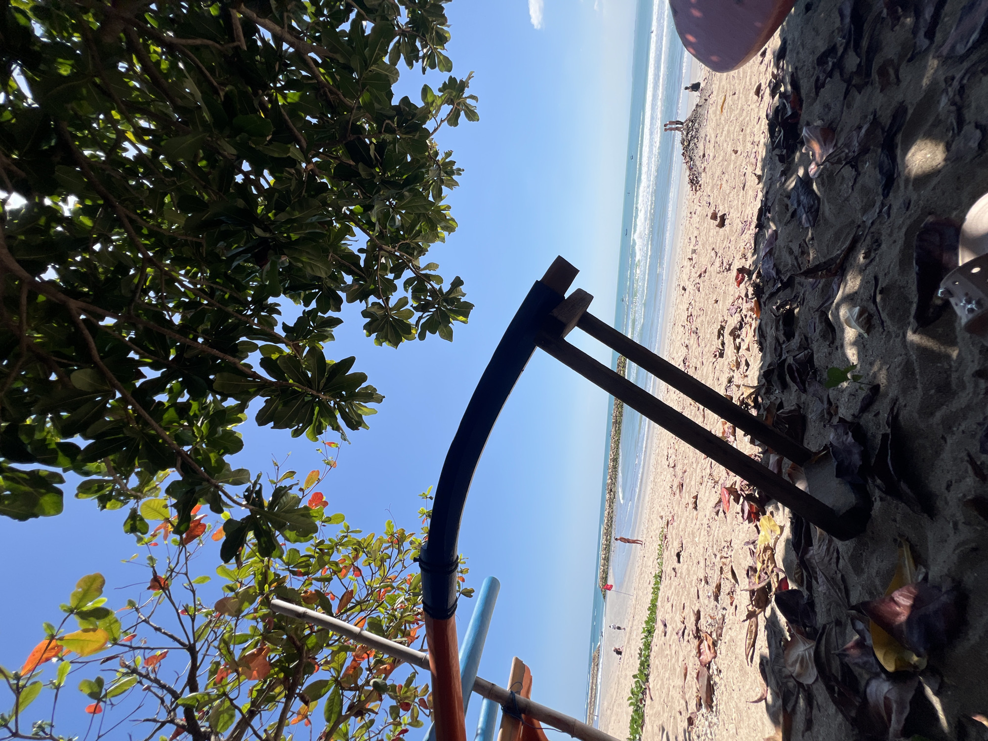 The image depicts a tranquil beach scene in Kuta, Indonesia. In the foreground, dry leaves and sand are scattered on the ground, with the shadow of a large tree providing shade. Two wooden posts, likely part of a beach chair or shelter, are visible supporting a dark, curved object. A colorful structure, possibly a part of a boat or beach equipment, is also present.
The mid-ground shows a sandy beach with a few scattered beachgoers. A red flag stands on the beach, and in the distance, a line of rocks forms a breakwater. The ocean is calm with gentle waves rolling towards the shore. Small boats can be seen on the water. The sky is a clear, vibrant blue, indicating a sunny day. The overall atmosphere is one of relaxation and natural beauty.