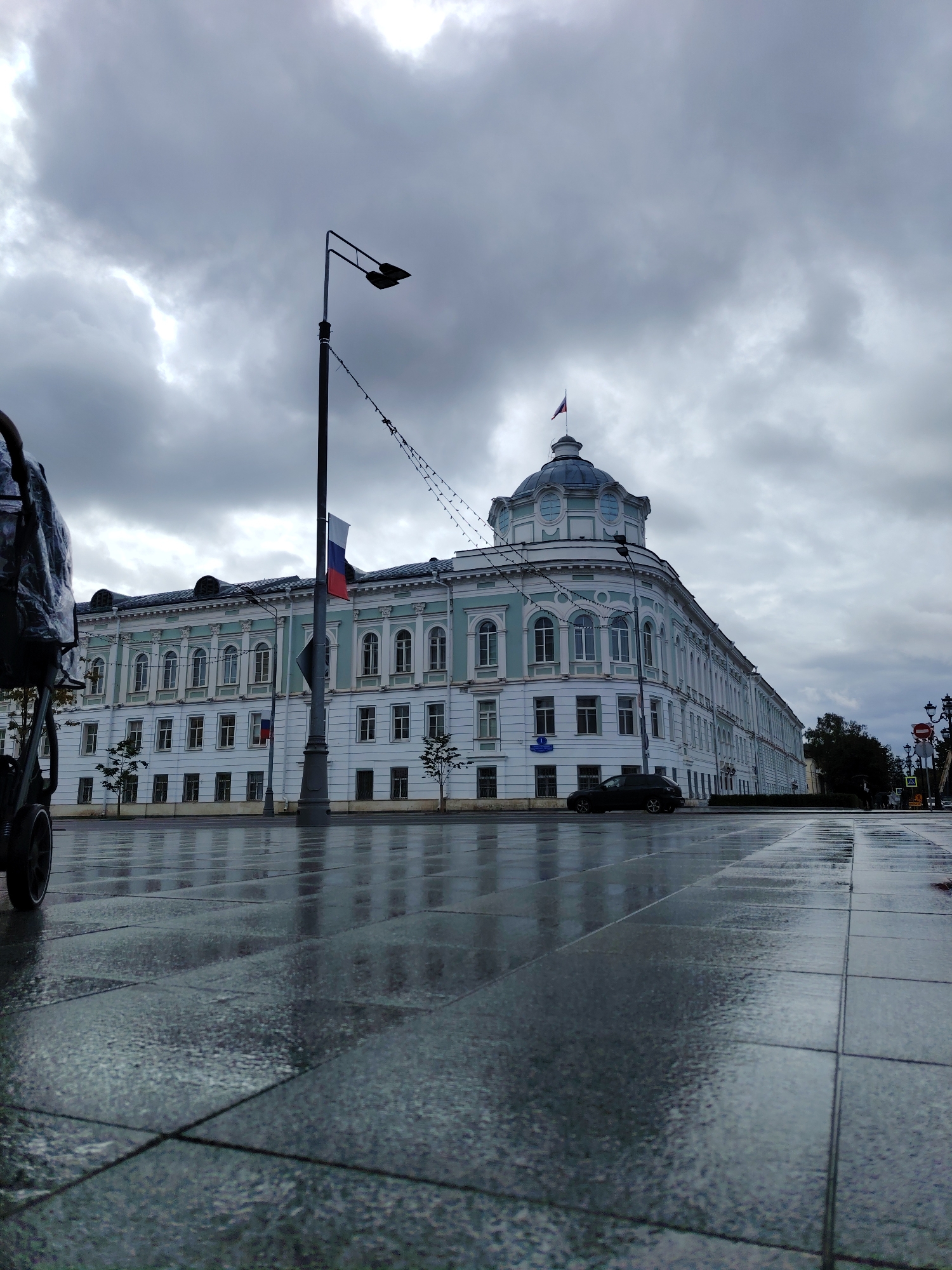 A low-angle, wide shot captures a modern Russian city scene on a cloudy day, likely in Tver, Russia. The foreground features a wet, tiled pedestrian walkway reflecting the overcast sky and surrounding architecture. To the left, the rear of a stroller or cart, partially covered in a gray plastic bag, is visible. A black car drives on a wet road adjacent to the walkway, with the building's reflection also visible on the wet road. In the middle ground, a large, light blue and white building with a classical architectural style, featuring numerous arched windows and decorative white trim, dominates the frame. A prominent cupola topped with a Russian flag crowns the building's corner. A tall, dark lamppost with two light fixtures stands in front of the building, adorned with a small Russian flag. In the background, more of the building stretches into the distance, and trees are visible. The sky is filled with dramatic, dark gray clouds, with patches of lighter sky suggesting breaks in the cloud cover.