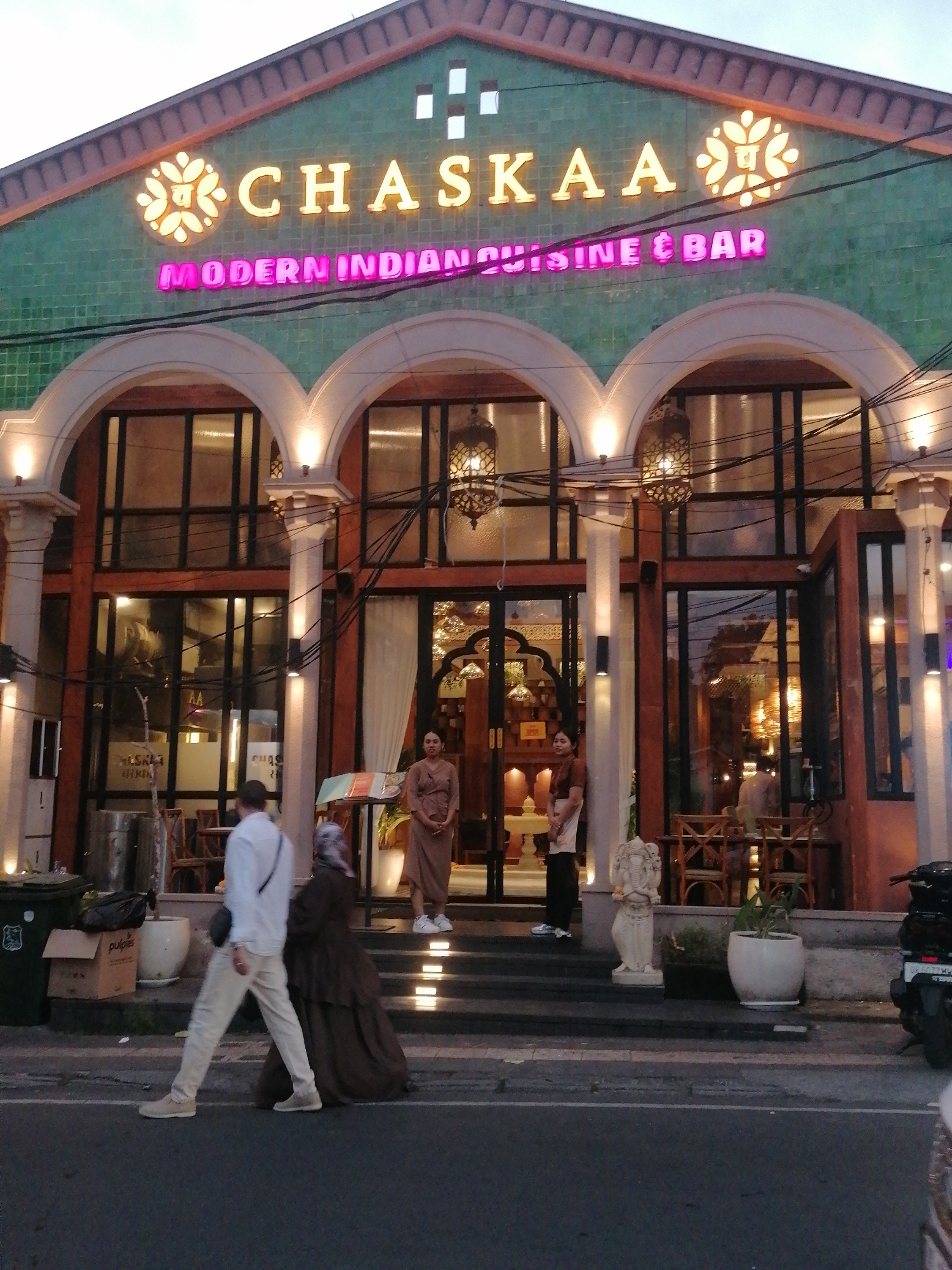 The image shows the exterior of a restaurant named "CHASKA A MODERN INDIAN CUISINE & BAR" located in the Ubud District, Indonesia. The restaurant has a green tiled facade with arched windows and is illuminated by neon signs. In front of the restaurant, several people are visible. Two women in traditional attire stand near the entrance, one to the left and one to the right. A man in a white shirt and beige pants walks past the entrance, followed by a woman in a dark dress. The street in front of the restaurant is paved, and there are plants and decorative statues on the side. A motorcycle is parked on the right side of the image. The overall scene suggests a commercial establishment operating in a public area.