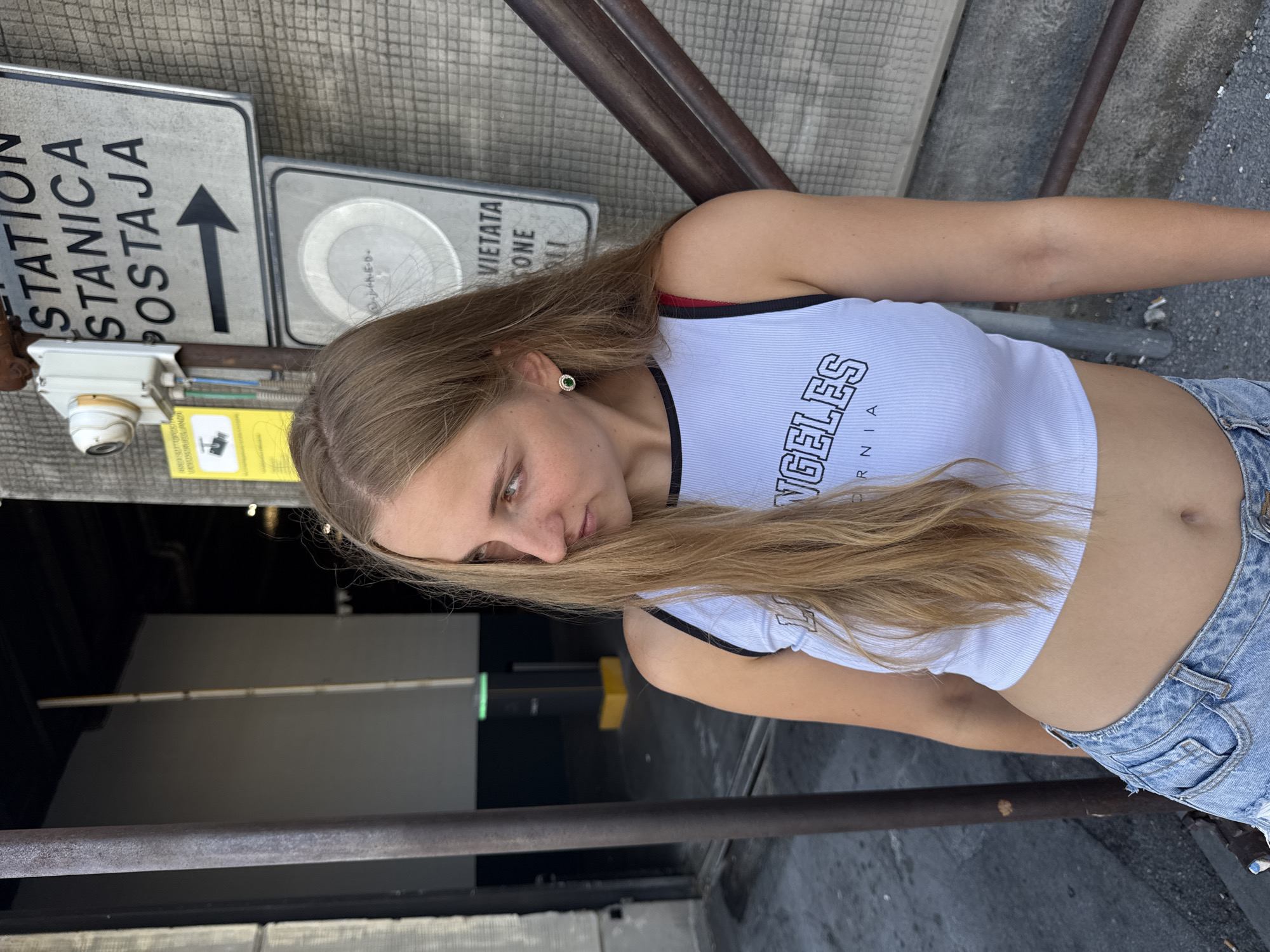 A young woman with long blonde hair is leaning against a metal railing next to a textured concrete wall. She is wearing a white ribbed crop top with "LOS ANGELES" printed on it and distressed denim shorts. She is looking to her left, and her right hand is resting on the railing. Behind her, a sign with the words "STATION STANICA POSTAJA" and an arrow pointing right is visible. There is also a security camera mounted above the sign. The environment appears to be an urban area, possibly a parking garage or an entryway to a building, with a dark interior visible in the background. A yellow and black bollard is also present in the background. The scene is likely in Trieste, Italy, as suggested by the multilingual signage.