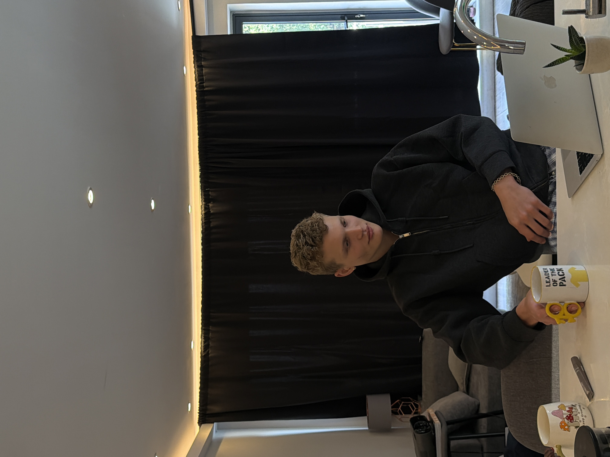 A young man with blonde, curly hair is seated at a white table. He is wearing a dark grey hooded sweatshirt and plaid pants. He is looking down and to his right. In front of him is a silver laptop, a white mug with a yellow handle that says "LEADER OF THE PACK" and has a yellow t-shirt graphic on it, and another mug with mushrooms and a brown pen. To his left, a portion of a sofa and a lamp are visible. Behind him, black curtains are drawn across a large window. The room appears to be a modern apartment in London, United Kingdom, likely a living room or kitchen area, with subtle recessed lighting in the ceiling.
