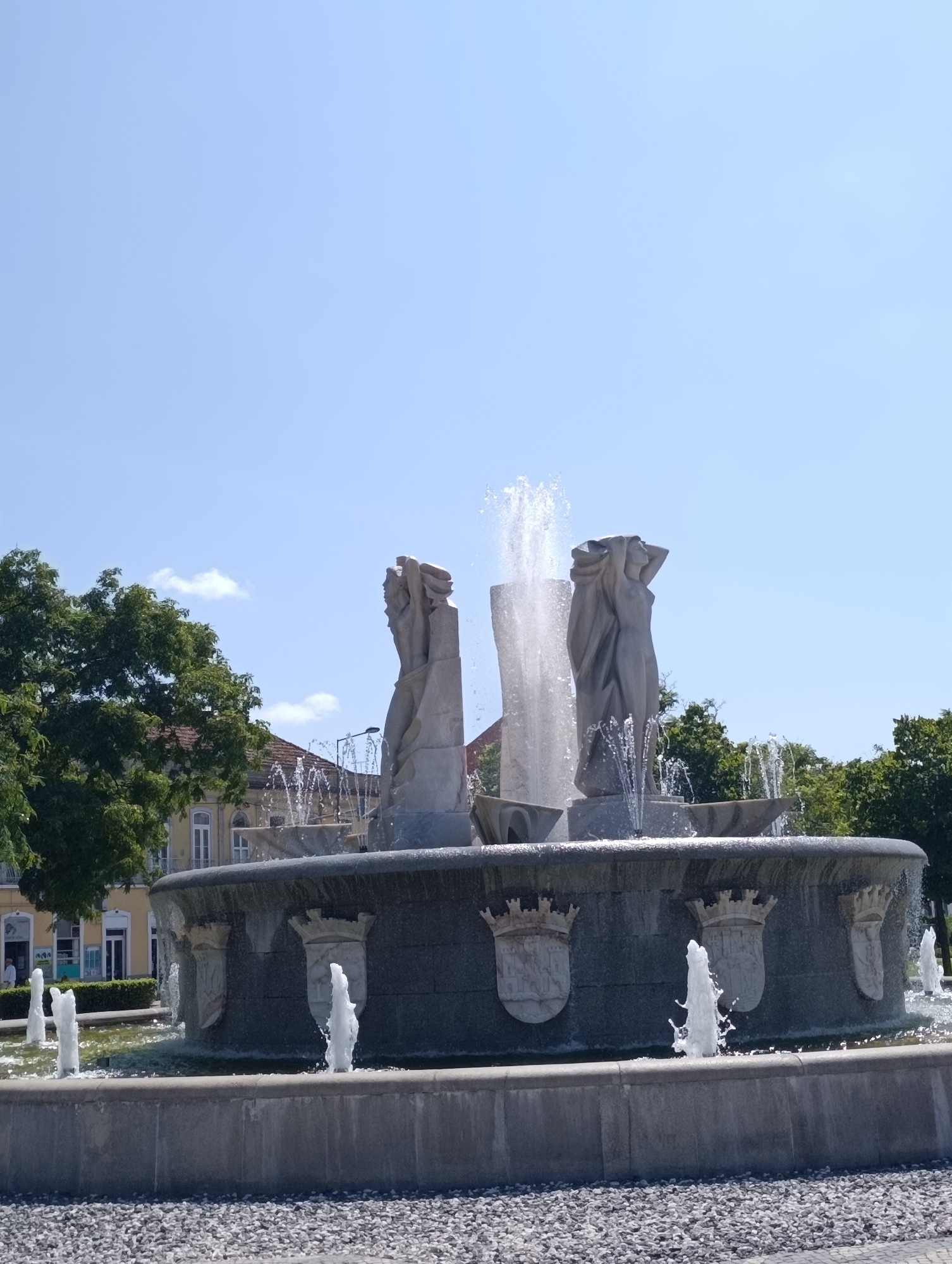 This is a daytime, eye-level, medium shot of the Monumental Fountain of the Discovery (Monumento à Descoberta) in Setúbal, Portugal. The fountain features a large, multi-tiered basin with several sculpted figures and water jets. Two prominent, abstract stone sculptures of human-like figures are centrally located, with water spraying upwards between them. The basin is decorated with shields bearing crowns. Smaller water jets are visible in the foreground and around the base of the fountain. Lush green trees line the background, with buildings with yellow facades and tiled roofs visible behind them. The sky is clear and blue with a few scattered white clouds. The ground in the foreground is covered in small, gray pebbles. The scene is brightly lit by natural sunlight.