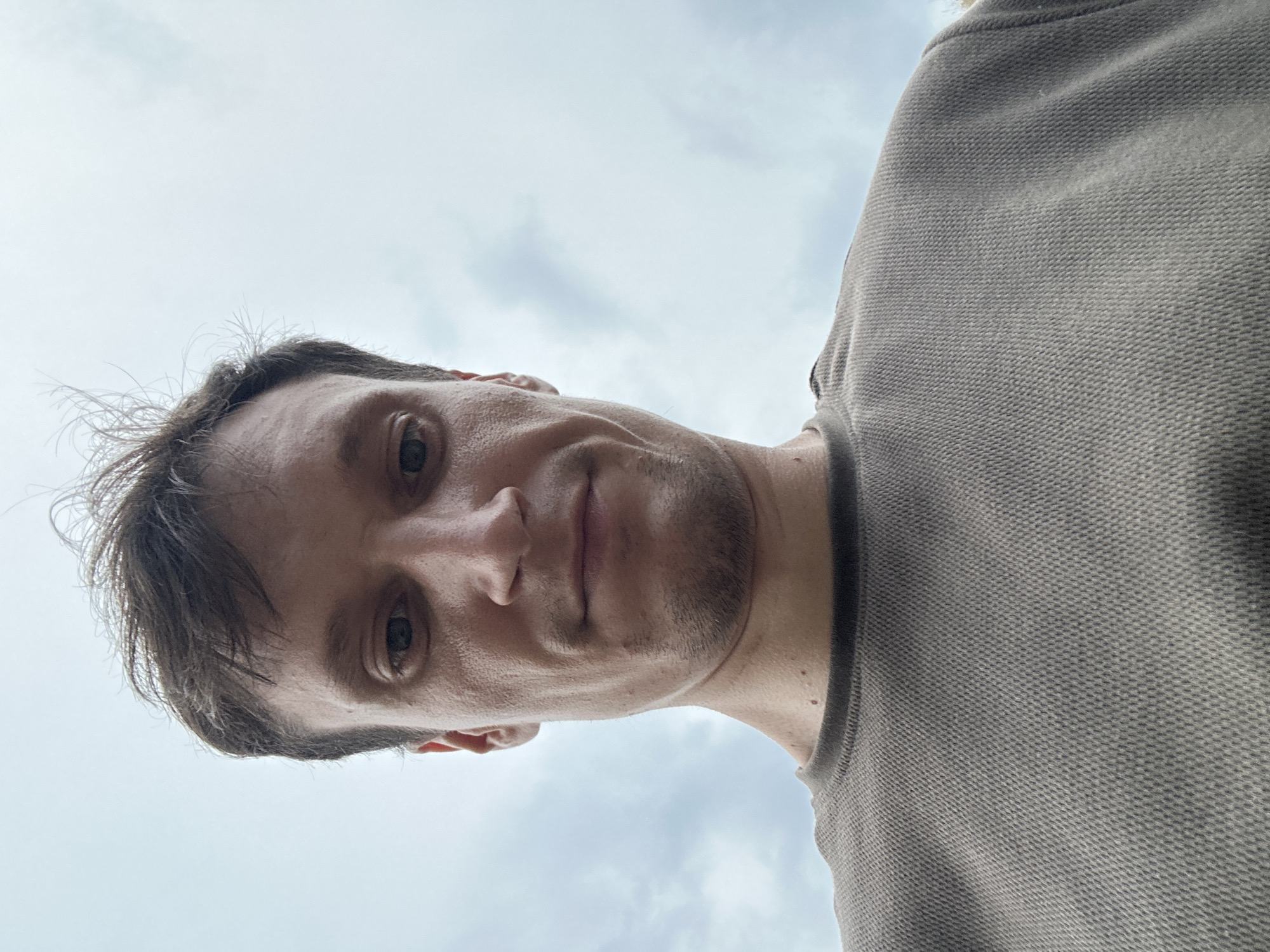 A low-angle, close-up, outdoor selfie captures a Caucasian male from the chest up against a cloudy sky. The man has short, brown, messy hair, dark eyes, and a neutral expression. He is wearing a grey, waffle-knit long-sleeved shirt. The image does not provide any specific visual cues to pinpoint its location within Kamianets-Podilskyi, Ukraine. The background consists solely of an overcast sky.