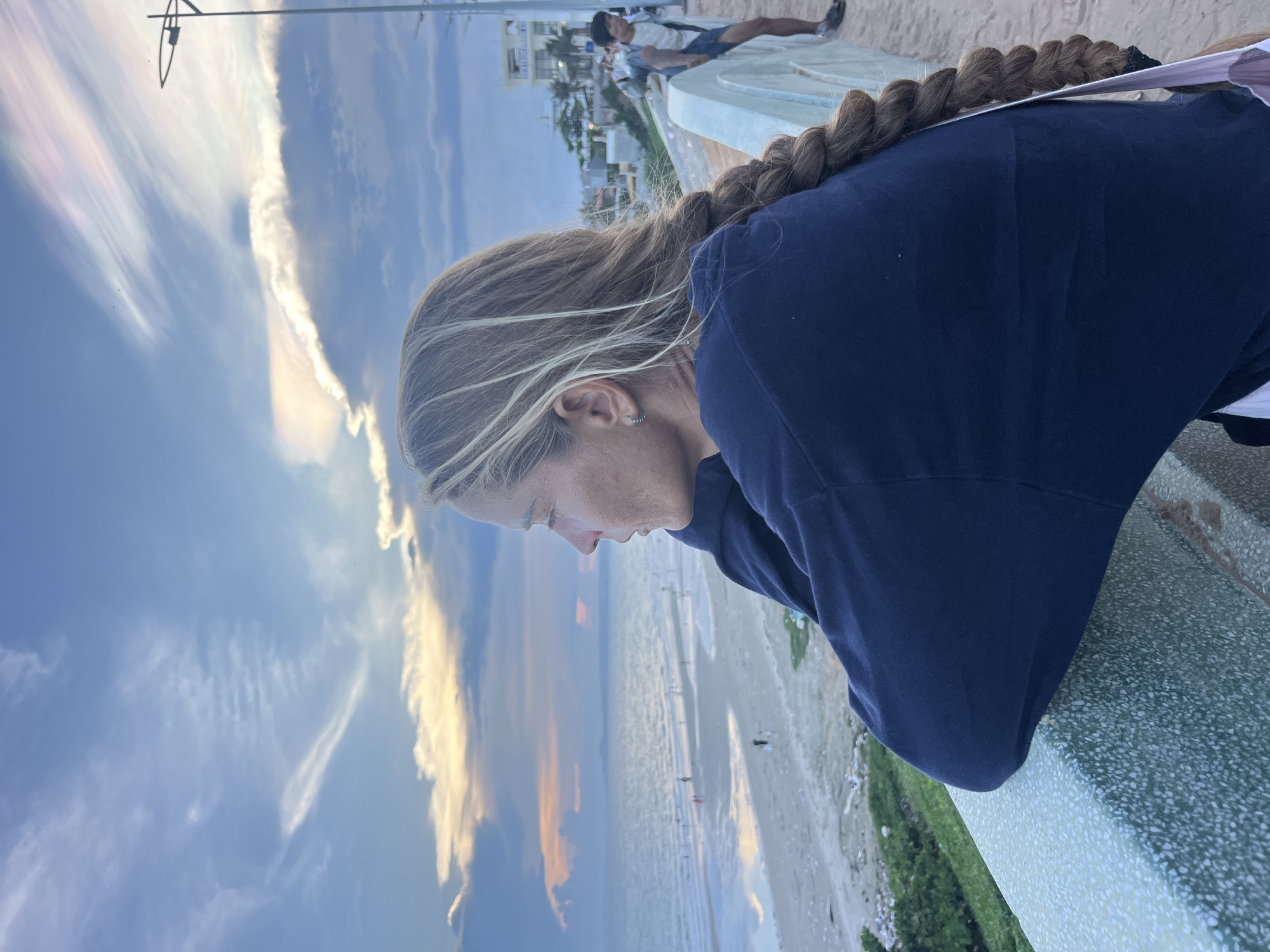 An outdoor, medium shot captures a woman from the shoulders up, facing left and looking out at a beach and the sea in Mũi Né, Vietnam. The woman has long, braided blonde hair that extends down her back. She is wearing a dark navy blue sweatshirt. Her left arm is resting on a textured, light green and white railing. In the background, the ocean stretches out, meeting a sandy beach with several small figures scattered across it. The sky above is a gradient of blues and oranges, with wispy clouds tinged with pink and yellow, indicating either sunrise or sunset. A streetlamp is visible on the right side of the frame, along with buildings and another person walking in the distance. The overall scene evokes a sense of calm observation of the coastal environment.