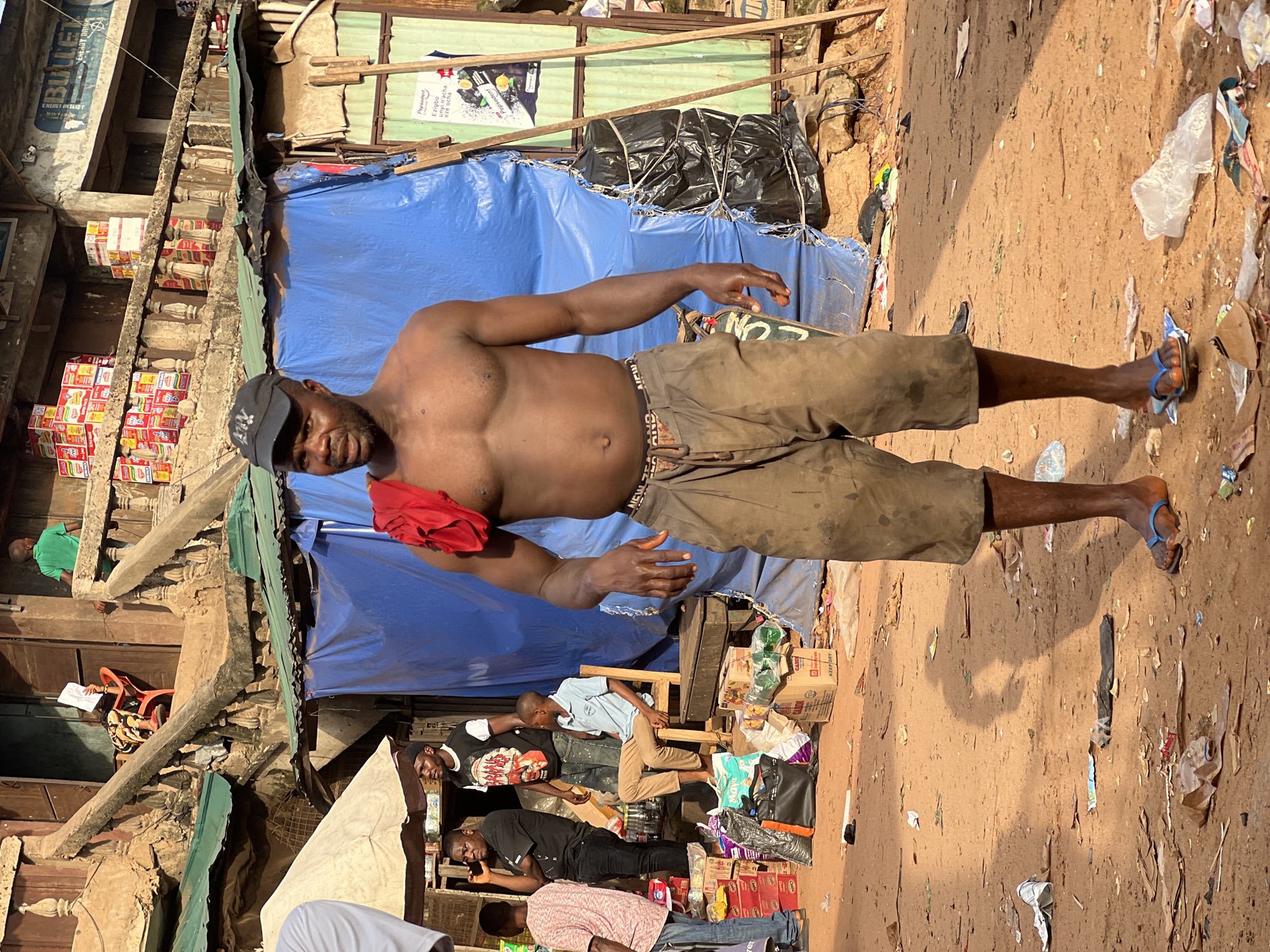 A man stands in a dusty, outdoor market, possibly Nkpor, Nigeria. He is shirtless, wearing a black baseball cap and baggy, dirt-stained tan shorts with a patterned waistband. A red cloth is draped over his left shoulder. His feet are bare, save for blue flip-flops. Behind him, a blue tarp is erected as a stall, with a stack of black garbage bags tied together to its right. To the left of the man, several people are visible, some engaged in conversation and others looking towards the camera. Scattered on the ground are various items including discarded wrappers, boxes, and plastic bags, indicative of a busy marketplace. The background features weathered buildings and more market stalls.