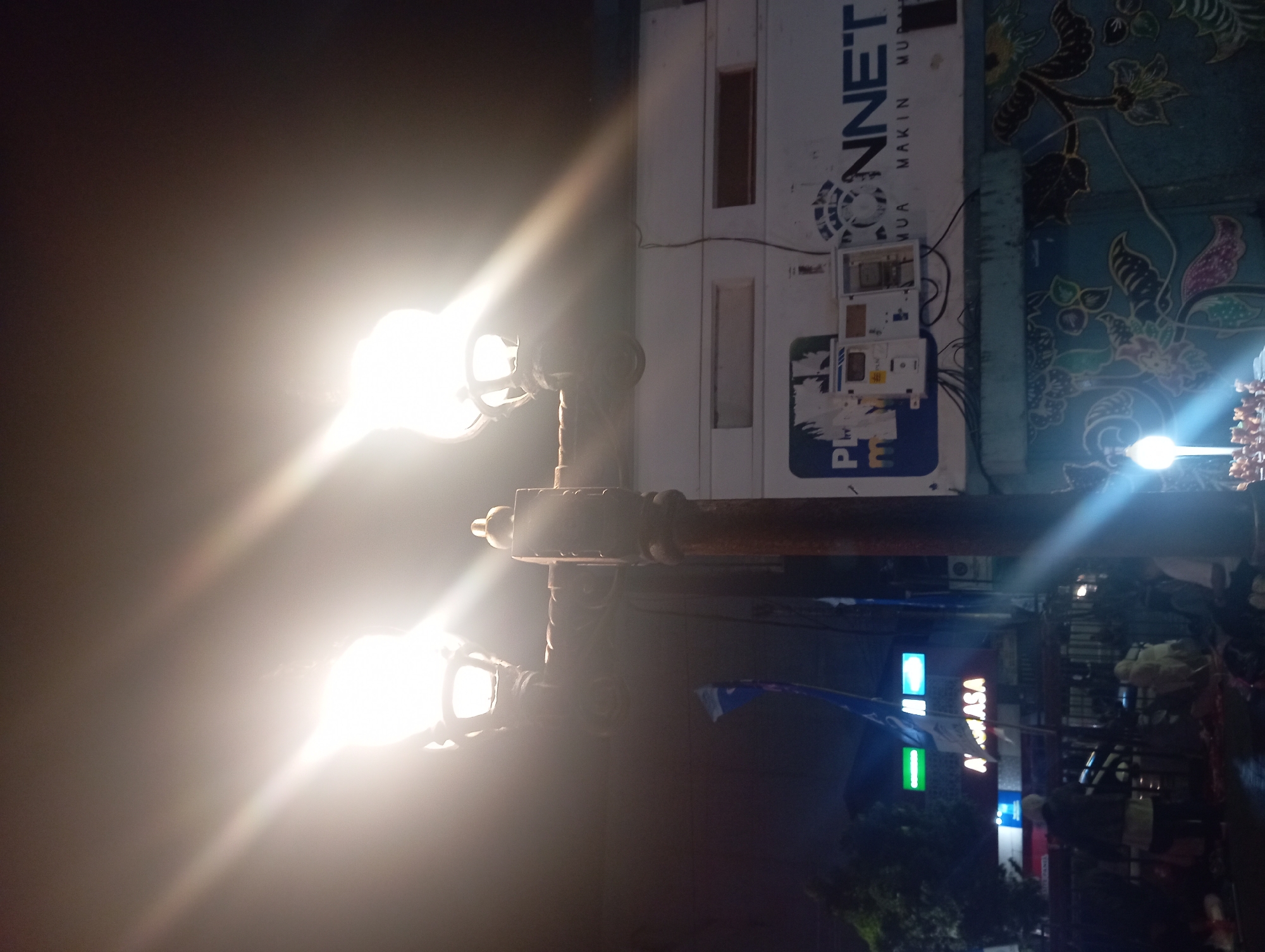 This is a nighttime street-level photograph taken in Purwokerto, Indonesia. The focal point of the image is a double streetlamp with two visible bulbs emitting bright light. The streetlamp is mounted on a dark, ornate pole. In the background, a white building is visible with various signs and electrical boxes mounted on its facade. One prominent sign reads "PHONNET" in large blue letters, accompanied by smaller text below. To the left, a sign with red and white lettering that appears to spell "ASA" is partially visible. Several flags, predominantly blue, are also present. The lower portion of the image shows the street with some greenery and what appear to be people and objects, possibly vendors or pedestrians, although they are indistinct due to the low light conditions. The scene suggests a commercial or public area at night.