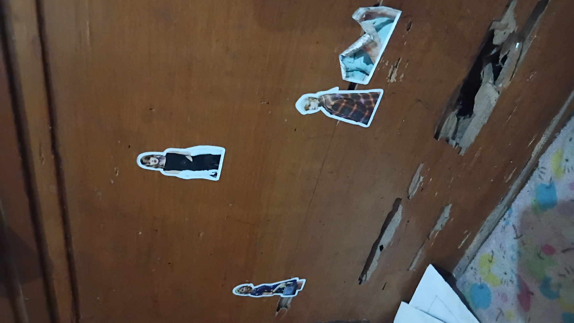 The image depicts a close-up view of a wooden surface, likely a piece of furniture, with several stickers of a woman in various poses. The wood shows signs of wear and damage, with several large gouges and chipped areas revealing the underlying material. One prominent gouge in the lower right quadrant is deep enough to show a dark interior. To the left of the damaged wood, a colorful, patterned fabric is partially visible, possibly a rug or blanket.
The stickers are applied to the wooden surface at different heights and positions. The topmost sticker shows a woman in a black dress with a white outline. To the left and slightly lower is another sticker of a woman in a blue dress, carrying a handbag. Further to the right and lower than the first sticker is one of a woman wearing a plaid jacket and a light-colored scarf. Near this sticker, to its right, are two more stickers that appear to be pieces of a larger image, possibly abstract or depicting a landscape, with prominent blue and white colors.
The lighting suggests an indoor environment, and the focus is on the surface of the wood and the stickers. There are no discernible elements in the image that would directly indicate a specific location within the city of Prabumulih, Indonesia. The scene is static and offers no information about actions or interactions.