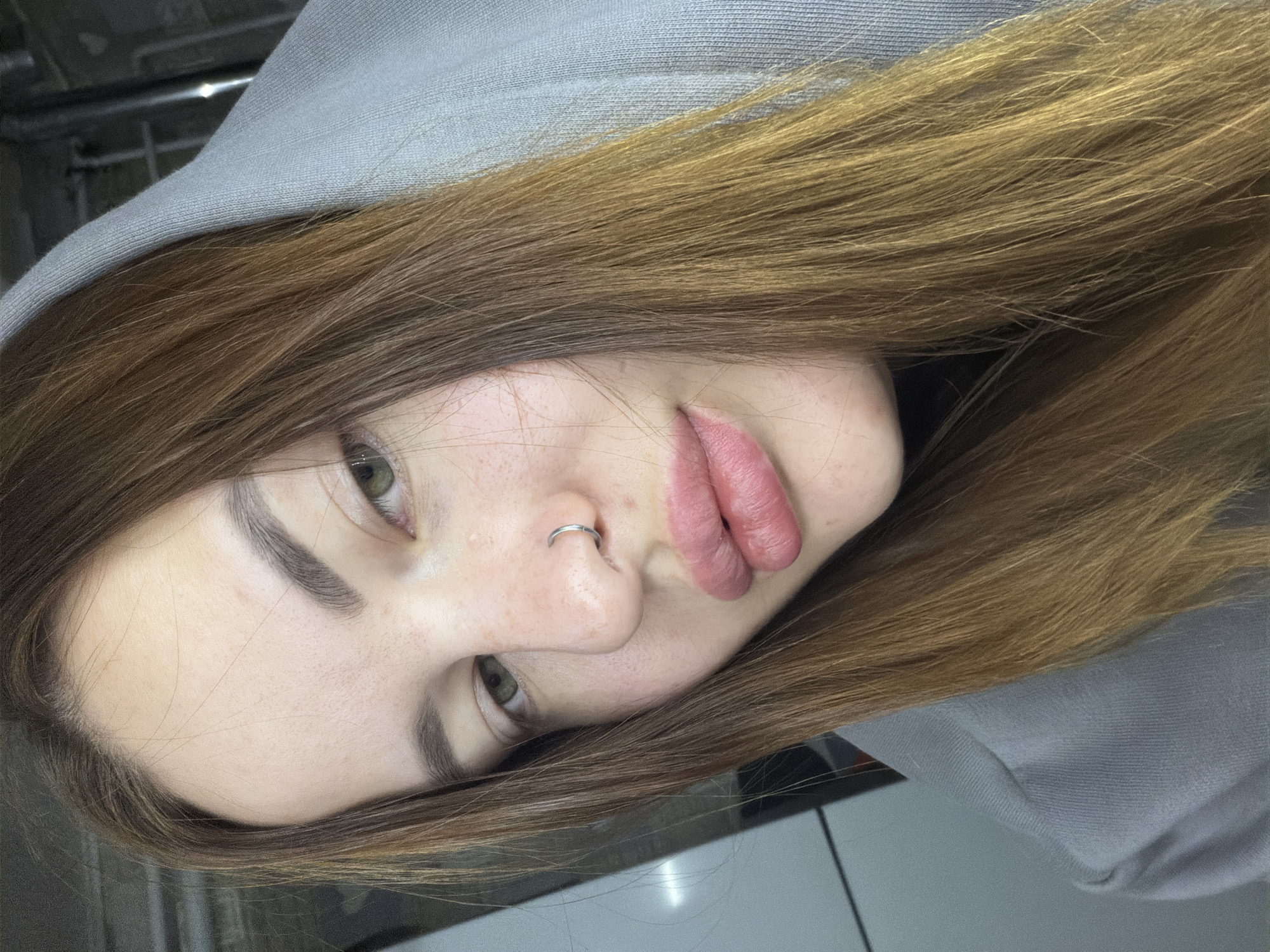 The image is a close-up portrait of a young woman, presented with her head tilted to the right due to the image's orientation. She has long, light brown to auburn hair that cascades around her face and over the shoulder of a light grey hooded sweatshirt she is wearing. Her eyes, appearing to be green or hazel, look directly towards the viewer with a neutral, perhaps slightly contemplative, expression. Her eyebrows are dark and well-defined, and her skin shows a scattering of freckles across her nose and cheeks. A silver ring septum piercing adorns her nose. Her lips are full, tinted with a soft pink or mauve color, and appear slightly parted. The background is dark, blurred, and indistinct, suggesting an indoor setting. Some metallic or structural elements can be faintly seen in the upper left corner. The image captures a posed moment with no specific activity taking place. There is no visible text. The location context is Port-Katon, Russia.
