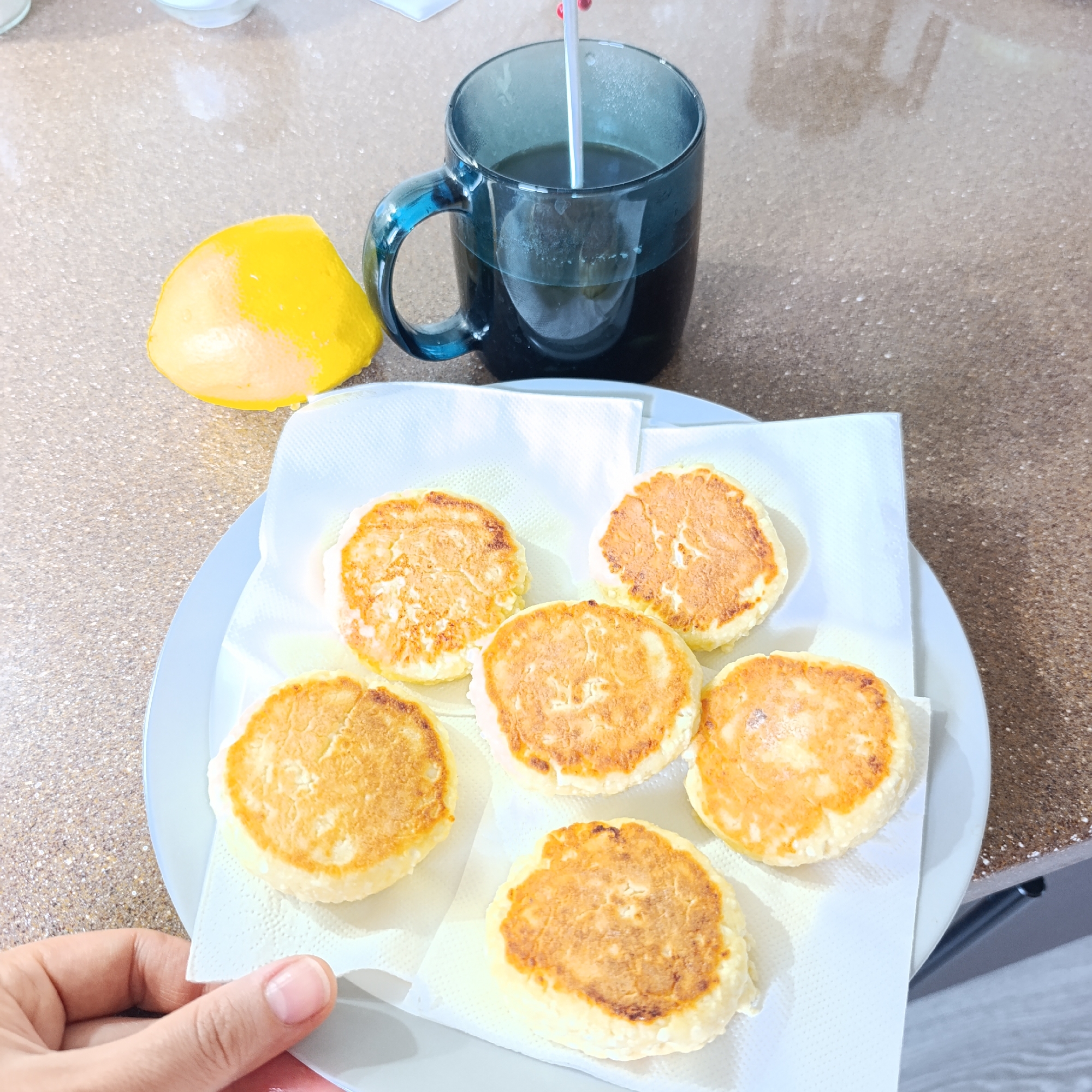 The scene depicts a hand holding a plate with six round, golden-brown patties placed on a white paper towel. A mug filled with a dark liquid and a stirring utensil stands nearby, along with a wedge of yellow fruit. The setting appears to be a kitchen or dining area, with a light-colored countertop visible. The location of Марковский, Russia, is not explicitly visible or inferred from the image.