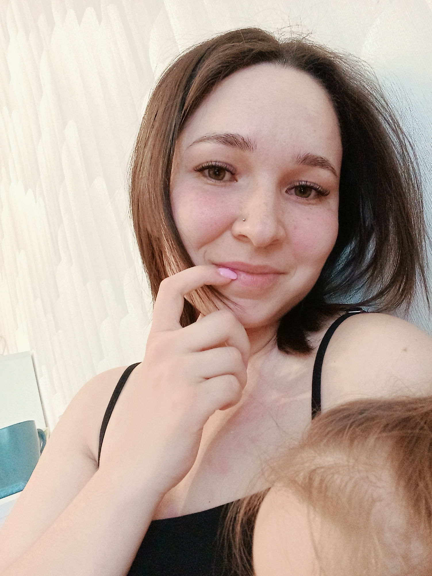 An indoor, eye-level photograph features a smiling adult female and a partially visible infant. The female, centered, possesses shoulder-length light brown hair, brown eyes, and a fair complexion with a visible nasal stud piercing on the left nostril. She is wearing a black, thin-strapped top. Her left index finger, with pink nail polish, touches her chin. She looks directly at the camera. In the lower right corner, the top of an infant's head, displaying light brown hair, is partially visible. The background consists of a light-colored wall with a textured, abstract, wavy vertical pattern. A light-colored object and a blue mug are partially visible in the left background. The specific location within Izhevsk, Russia, is not ascertainable from the visual evidence.