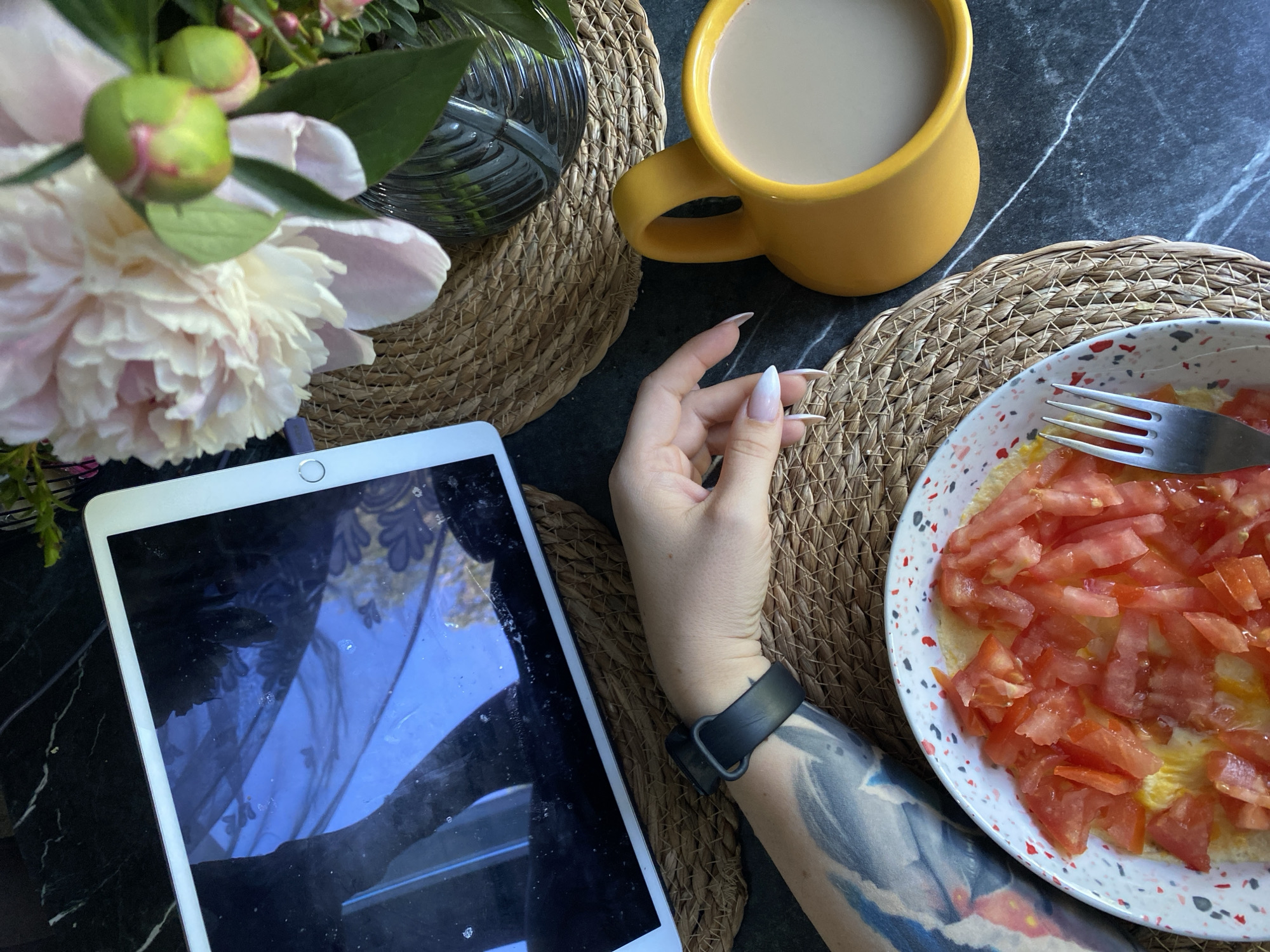 The image depicts a top-down view of a breakfast setting on a dark marble countertop. In the foreground, a plate of scrambled eggs topped with diced tomatoes is visible, with a fork resting on the edge of the plate. To the right, a yellow mug filled with a light-colored beverage, likely coffee or tea, sits on a woven placemat. A woman's arm, adorned with a tattoo and a black smartwatch, extends from the left side of the frame, with her hand positioned near the plate. Further back, a white tablet is placed on another woven placemat. To the right of the tablet, a bouquet of peonies in various stages of bloom, including buds, is displayed in a glass vase. The scene is set indoors, with reflections of what appears to be curtains and possibly an outdoor scene visible on the tablet screen. There is no information in the image to suggest the location is in Chaikovsky, Russia.