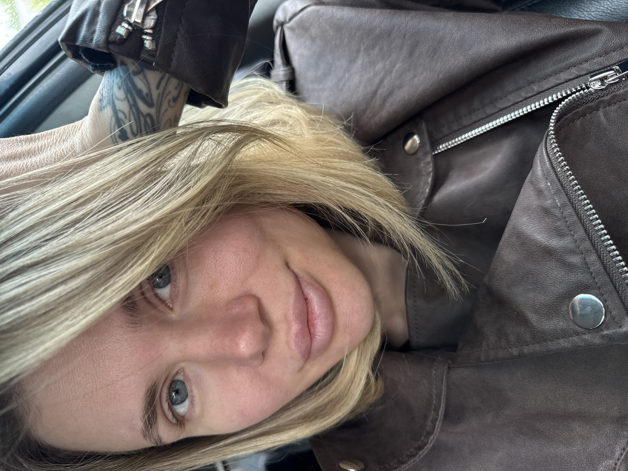 A woman with blond hair, blue eyes, wearing a brown leather jacket with silver hardware, is indoors. She has a tattoo on her arm. The location is unknown and it is not possible to ascertain if the location is in Gorno-Altaysk, Russia.
