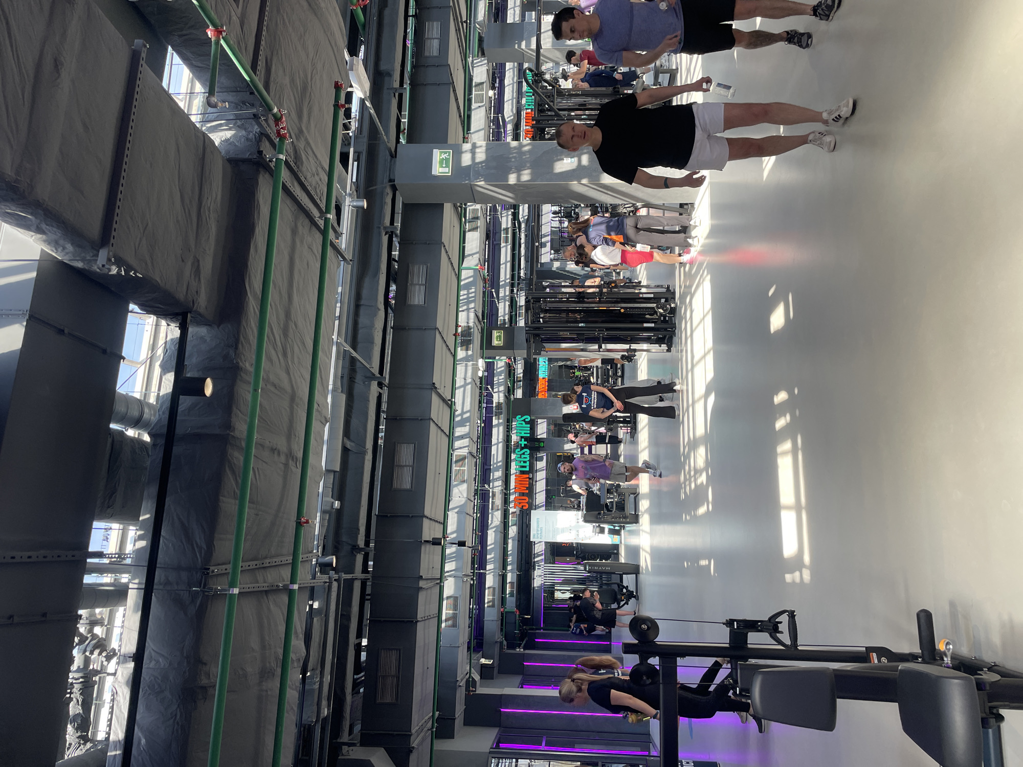 The image depicts the interior of a gym. Several people are engaged in exercise activities using various machines and equipment. The gym has a modern design with high ceilings and visible industrial ductwork. The environment is brightly lit with natural light coming through the windows. The location is Kazan, Russia.
