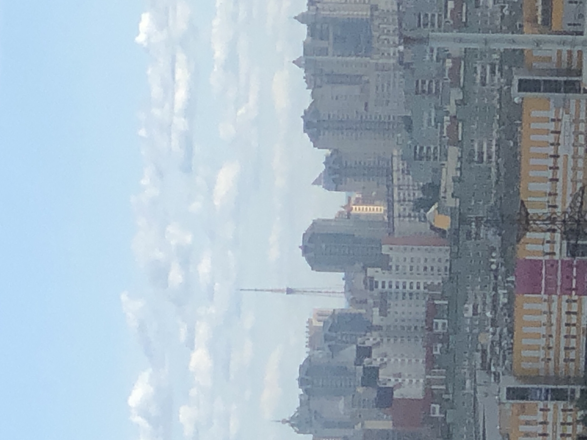 The image depicts a cityscape with numerous buildings of varying heights and architectural styles. The perspective is from a high vantage point, looking down and across the urban landscape. The buildings are densely packed, suggesting a major city. Some structures are modern high-rises, while others appear to be older, more traditional buildings. There is a prominent tower or antenna visible in the distance, piercing the sky above the rooftops. The sky is partially filled with clouds, creating a soft, diffused light, indicative of daytime. The overall color palette is muted, with grays, whites, and some hints of yellow and pink from the building facades. There is no visible text in the image. The scene conveys a sense of urban sprawl and the dynamic nature of a city. The activity taking place is the visual representation of city life, with buildings forming the infrastructure of daily existence. The time of day appears to be daytime, given the brightness of the sky and the ambient light. The weather seems to be partly cloudy.