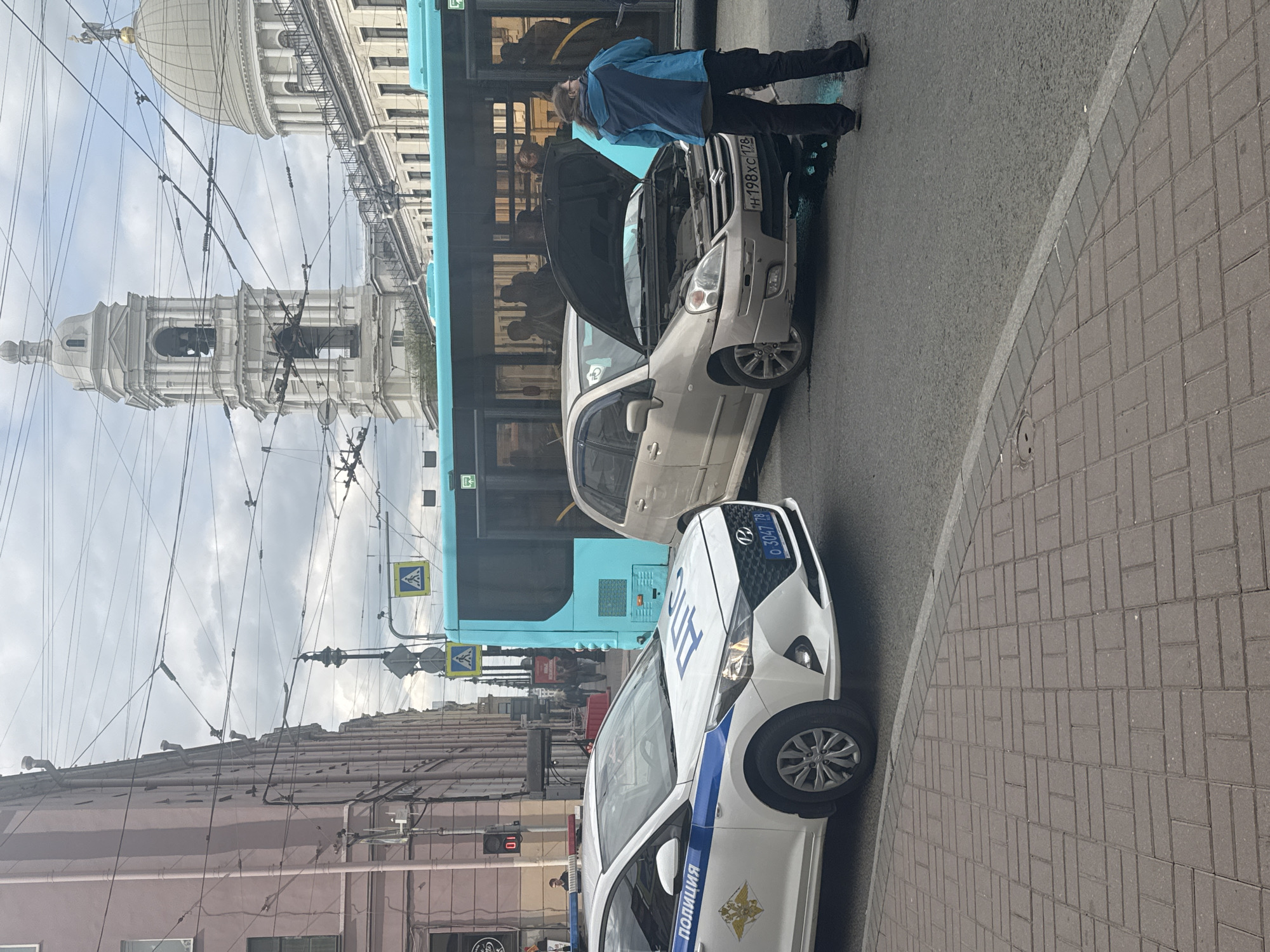The scene depicts a street in Saint Petersburg, Russia, where a traffic incident appears to have occurred. A light-colored car has its hood open, with a person in a blue jacket standing nearby. A blue bus is positioned directly behind the car, obscuring part of the view. A police car is parked in front of the light-colored car. The environment includes overhead electrical cables and a view of a prominent building with a dome, identifying the location as Saint Petersburg. The street is paved with cobblestones and there is a pedestrian crossing in view. A pedestrian traffic light is also visible.