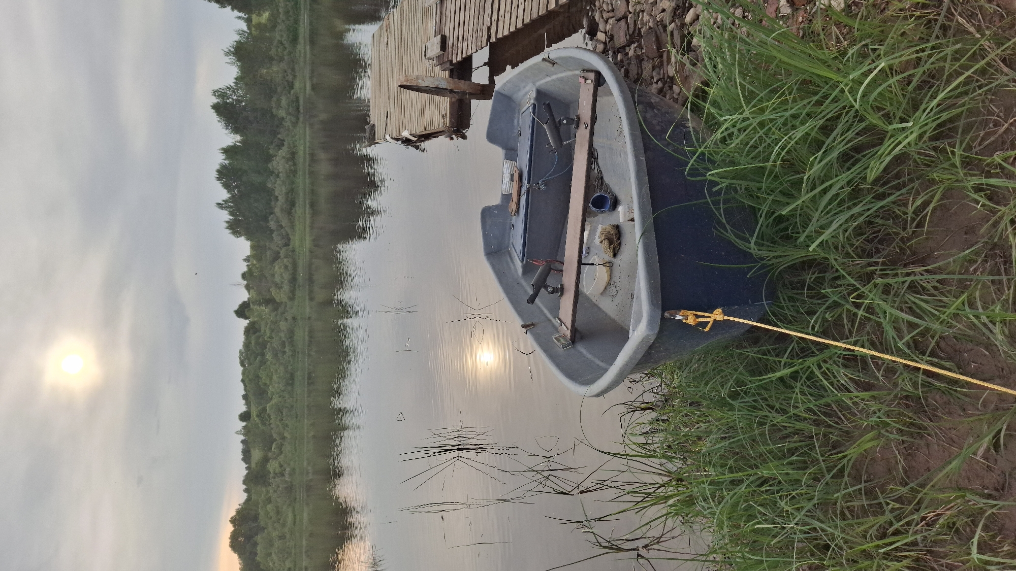 The image depicts a small, dark-colored boat moored on the shore of a calm body of water, likely a lake or river, under an overcast sky with the sun partially visible. The boat is a plastic fishing boat with two seats and two rod holders mounted on a wooden plank. A yellow rope is tied to the bow of the boat and extends towards the viewer, anchoring it to the shore. The shore is covered with green grass and some rocky patches. To the right of the boat, a wooden pier extends into the water. The background features a dense line of trees on the opposite bank of the water. The overall scene suggests a quiet, natural environment, possibly in the rural area of Sitno, Russia, given the typical landscape and rustic elements. There are no people visible in the image.