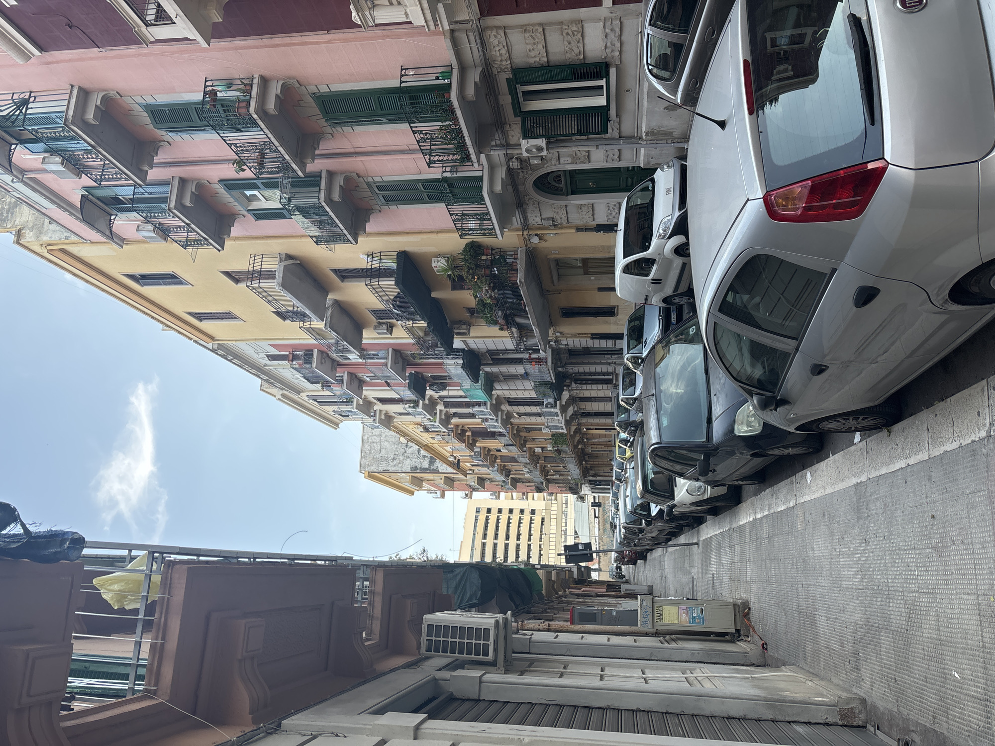 The image depicts a narrow urban street lined with multi-story residential buildings under a bright sky. The perspective looks down the street, with buildings on both sides and a line of parked cars on the right.
The buildings vary in color, featuring facades of pink, yellow, beige, and grey. Many have traditional dark green wooden shutters on their windows and numerous balconies with intricate wrought-iron railings. Some balconies are decorated with potted plants, and several air conditioning units are mounted on the building exteriors. In the distance, a taller, more modern building with many windows is visible.
Along the right side of the street, a series of cars are parked bumper-to-bumper. The closest vehicle is a silver hatchback, identifiable by the "FIAT" logo on its rear. Further along the street, a white commercial van can also be seen. The sidewalk or pavement on the right is a grey, textured surface.
The sky above is clear and light blue with a few scattered, wispy white clouds, indicating fair weather. The bright natural light suggests it is daytime, likely morning or afternoon. No people are visible in the scene, conveying a sense of quiet urban life.
Besides the "FIAT" logo on the car, a small, faded poster or sticker is present on a utility box or panel on the right sidewalk. While most of the text is illegible due to blurriness, "24H" and "HA" are partially discernible.