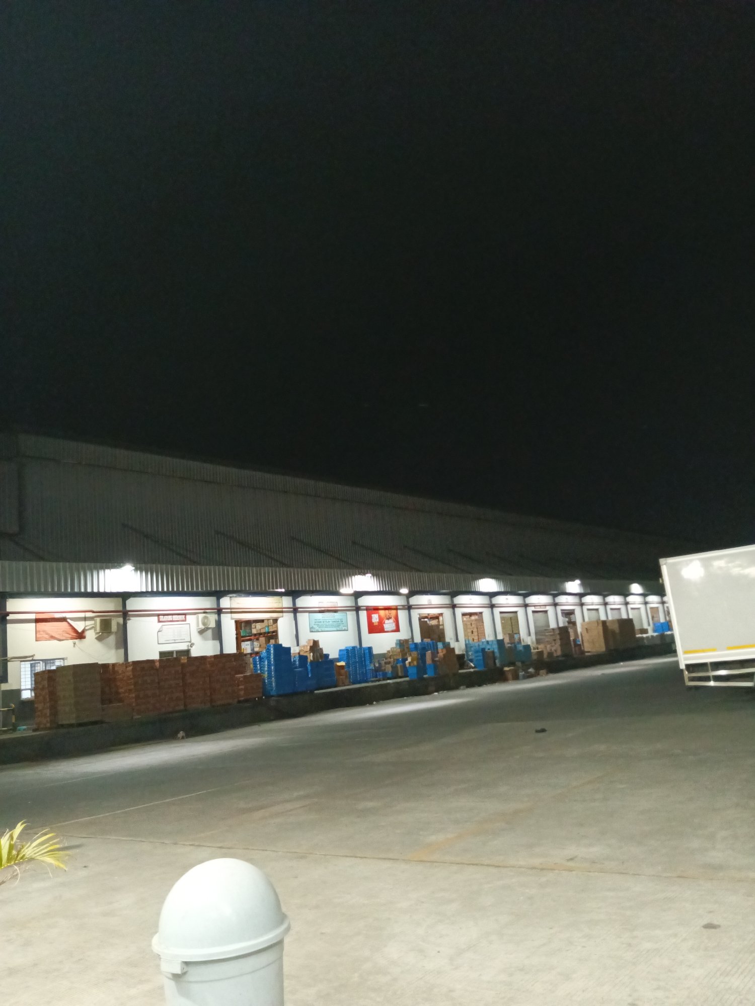 The image shows a loading dock area outside a warehouse at night. The warehouse has a long, corrugated metal roof supported by pillars, and a row of loading bay doors. Numerous boxes and pallets are stacked in front of the warehouse, some wrapped in plastic and some in blue plastic crates. A white truck is partially visible on the right side of the image. In the foreground, there is a white trash can and some yellow vegetation. The ground is paved concrete. There are no people visible in the image. The scene suggests a commercial or industrial setting, likely a logistics or distribution center. Without further information, it is not possible to confirm the specific location within Makassar, Indonesia.
