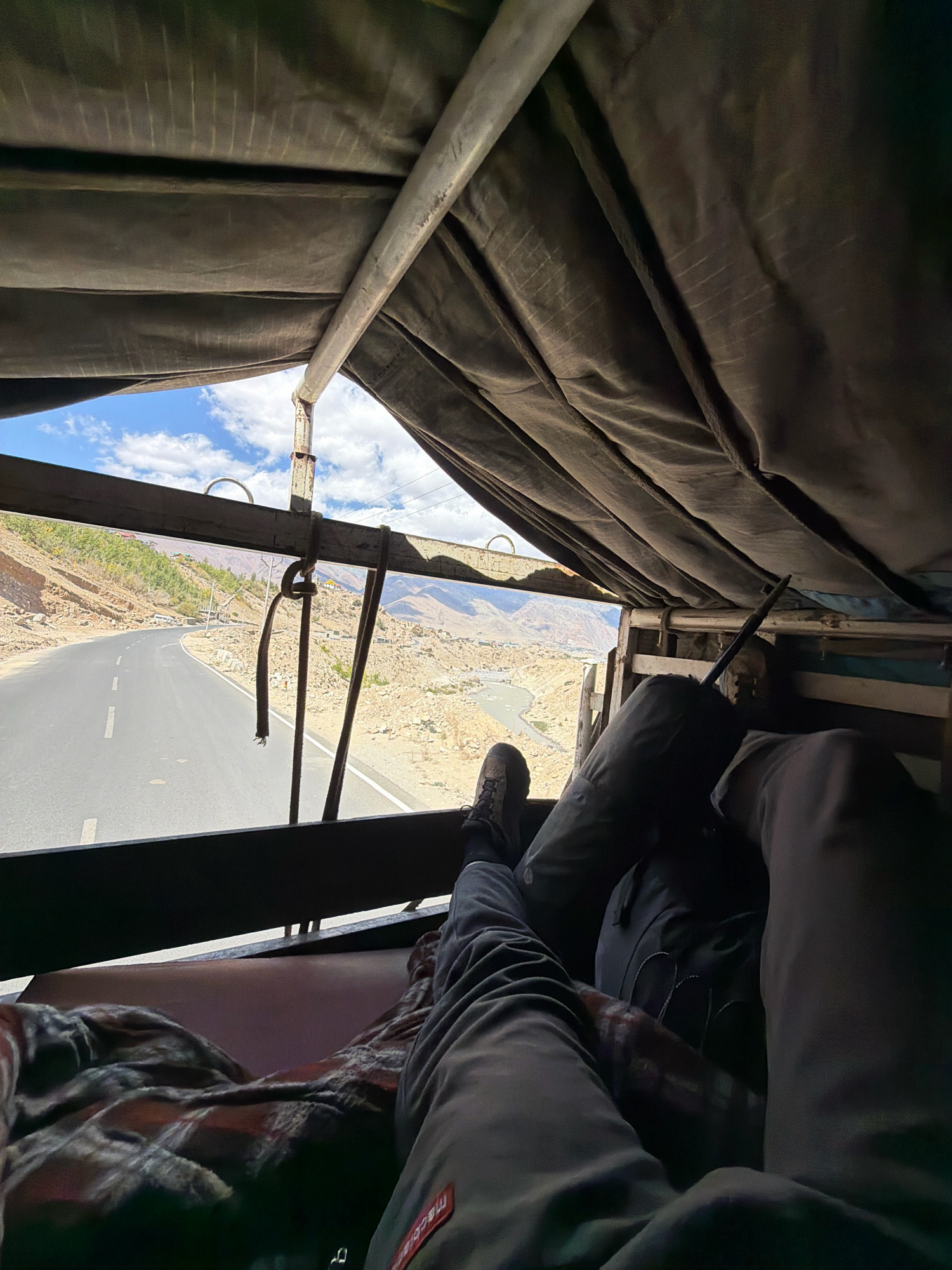 The image shows a view from inside a vehicle, likely a truck or utility vehicle, looking out through an open side. The foreground is dominated by the legs and feet of two individuals, one with a red and black plaid blanket draped over their lap, and the other wearing dark pants and hiking boots. The interior of the vehicle is covered by a dark, textured canvas material.
Through the opening, a paved road curves to the left, bordered by a barren, mountainous landscape with patches of green vegetation on the slopes. A river or body of water is visible in the valley below the road. The sky is blue with scattered white clouds. The scene suggests a journey through a mountainous region, possibly in the Himalayas. While the prompt mentions Padum, India, the image itself does not contain explicit visual cues to definitively confirm this location. However, the arid, mountainous terrain with a winding road and scattered settlements is consistent with such a region.