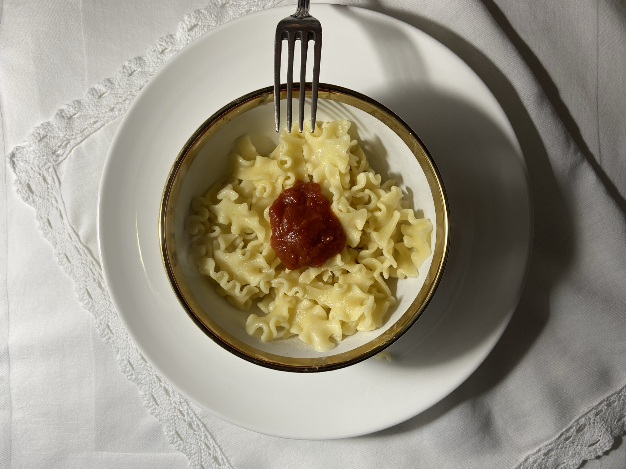 A close-up shot reveals a small portion of wavy pasta topped with a dollop of red sauce, served in a white bowl with a gold rim. The bowl sits on a larger white plate, which is placed on a white linen cloth with a delicate lace trim. A silver fork is stabbed into the pasta. The environment appears to be indoors with soft, diffused lighting creating gentle shadows. There is no indication in the image that this is in Budva, Montenegro.