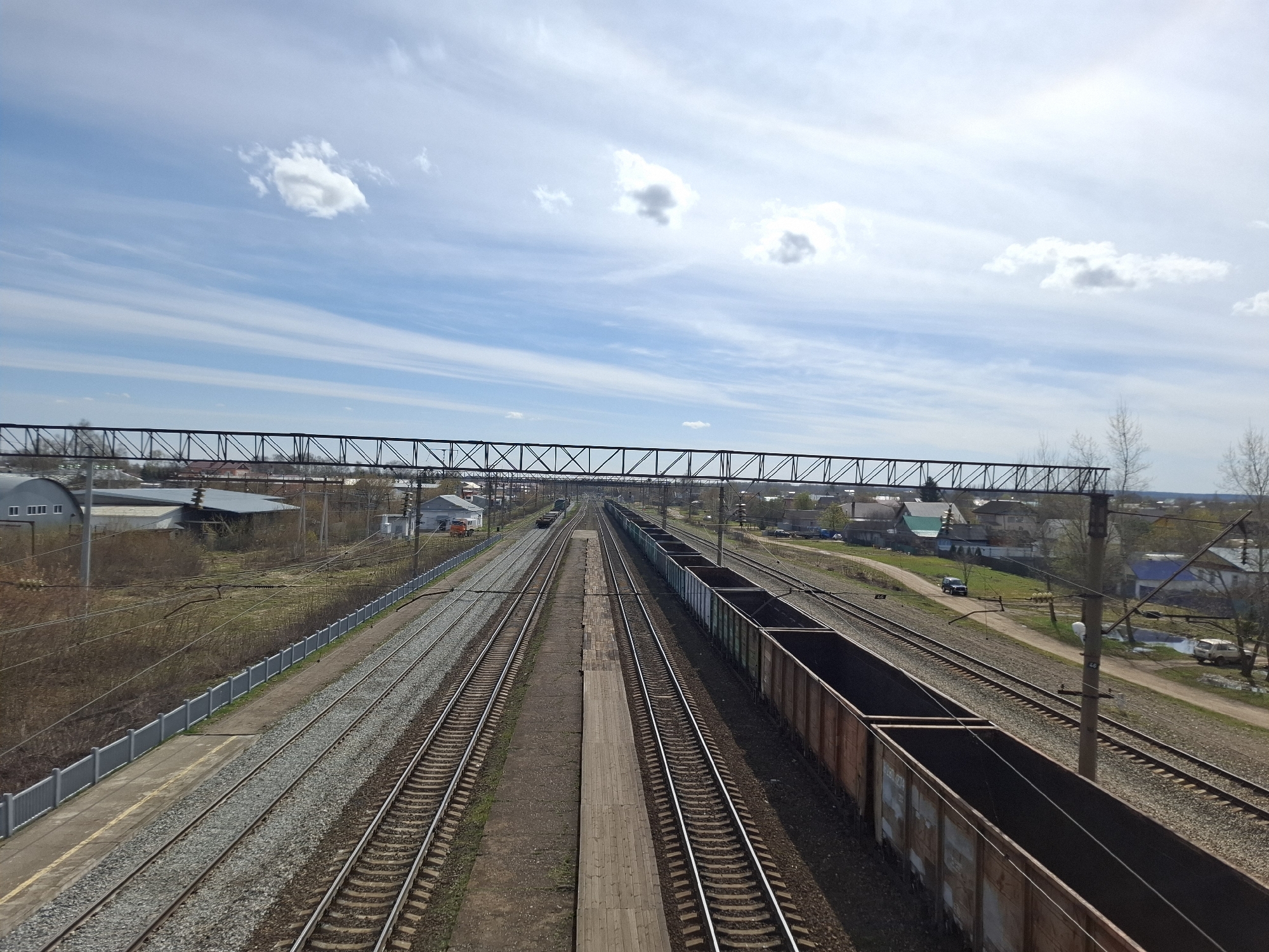 The image depicts a view of railway tracks in Амзя, Russia. The perspective is from an elevated position, possibly a bridge. Several parallel tracks run towards the horizon. Freight cars are visible on the rightmost track. A pedestrian platform is situated between the central tracks. A railway electrification system (catenary) is visible above the tracks. Residential buildings and vegetation line the tracks. The sky is blue with scattered clouds.