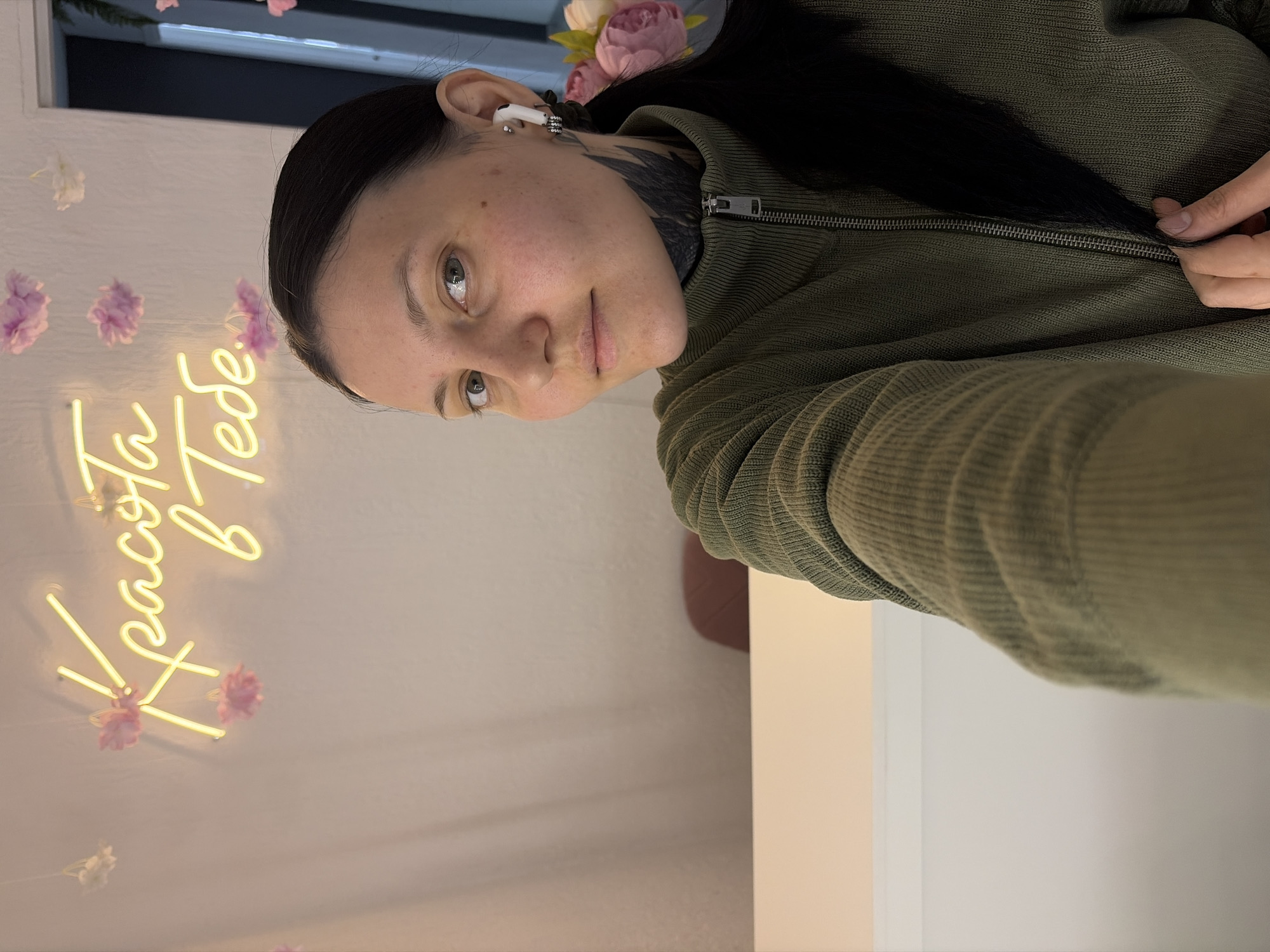A woman is in what appears to be an indoor setting. She is fair-skinned with dark hair and visible neck tattoos. She wears a green, zip-up sweater. An earbud is visible in her left ear. Behind her, there is a wall with floral decorations and a neon sign in Russian that says "Красота в тебе" (Beauty is in you). The location is not specified as the city of Chaikovsky, Russia in the image. The photograph appears to be a selfie.