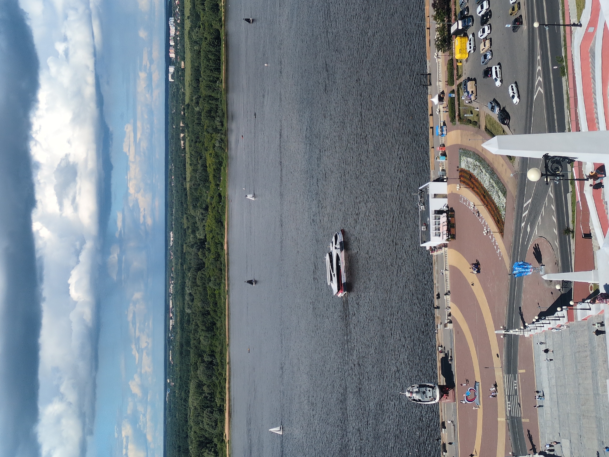 The image captures a wide, panoramic view of a riverfront area, likely on a sunny day with scattered clouds. The scene is divided into distinct sections: the expansive, dark grey water of a river on the left, a dense green treeline bordering the river, and a paved urban area with roads, walkways, and buildings on the right.
Several boats are visible on the water. One prominent vessel is a white and red catamaran-style boat moving through the river. Other smaller sailboats, some with dark sails and others white, are also scattered across the water, suggesting a recreational or sporting event.
The urban area features a road with parked cars, including a distinctive bright yellow vehicle. Pedestrians are seen walking along the waterfront promenade, which is paved with decorative patterns of yellow and red. There are also buildings visible along the edge of the treeline and further back in the distance, some appearing to be residential or commercial. A large blue parachute or banner is set up on the paved area near the water's edge. The presence of streetlights and people suggests it is daytime.
The overall impression is a vibrant, active waterfront scene, combining natural elements of the river and trees with urban infrastructure and human activity. The time of day appears to be late morning or afternoon, judging by the lighting and shadows. There is no visible text in the image.