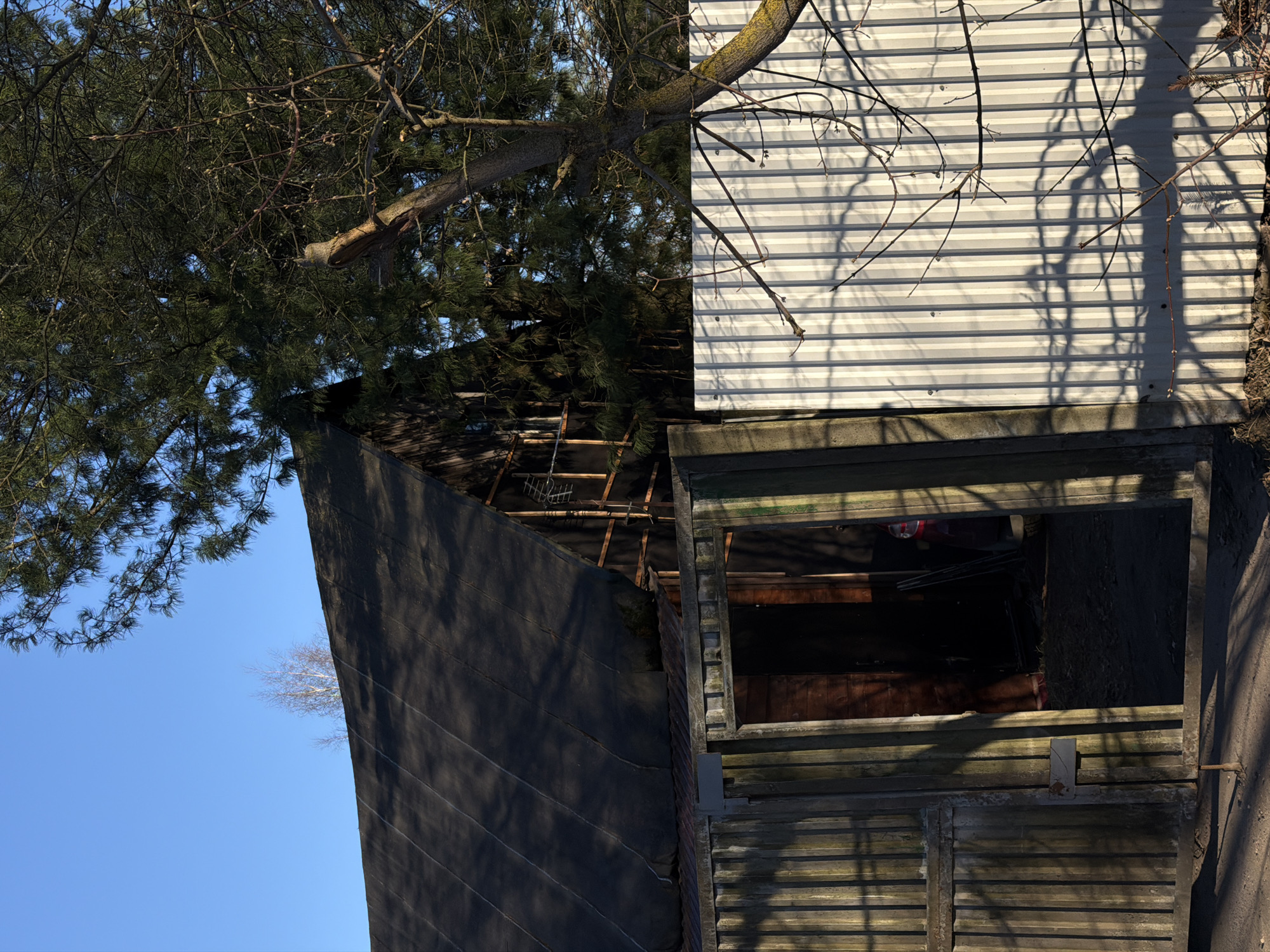 An exterior view shows a ramshackle wooden structure, potentially a shed, with an open window, set against a backdrop of blue sky and tree branches, and what appears to be the exterior siding of a building with horizontal lines. The setting seems to be in a rural or semi-rural area. There is no evidence in the image to confirm this is in Lavriki, Russia.