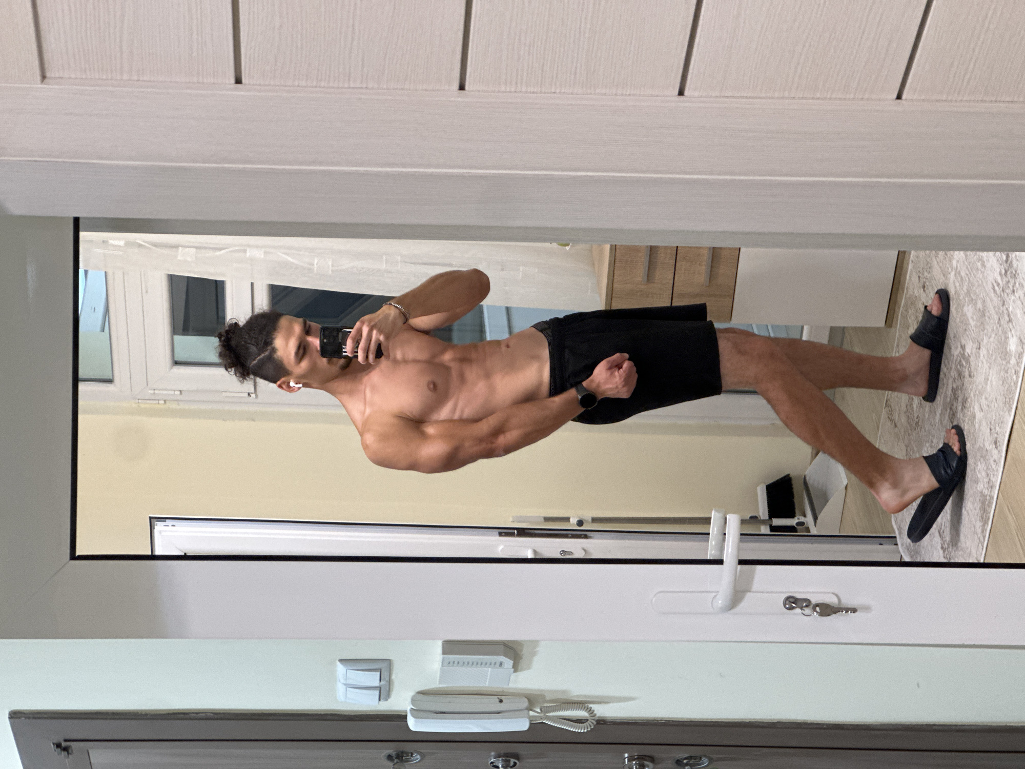 A young adult male is standing in what appears to be a hallway, taking a selfie in a mirror. He has a muscular build and is wearing black shorts and black slide sandals. He has his hair pulled up in a bun. In the foreground, to the left, is a door with a key and a security intercom system mounted on the wall next to it. To the right of the male subject, another door is visible, which is slightly ajar, revealing a glimpse of a room with wooden drawers and a window with sheer curtains. A broom and dustpan are visible in the reflection of the mirror in the background. The floor is covered with a rug. The overall setting suggests an indoor residential environment, possibly an apartment. There is no information to ascertain the location in Varna, Bulgaria.