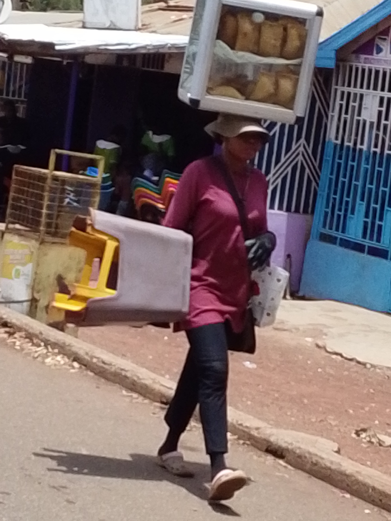 A street scene shows a person carrying items on their head and in their arms. The individual is wearing a hat, a maroon shirt, dark pants, and slip-on shoes. They are walking on a paved road near a curb. On their head, they balance a glass-fronted box filled with what appears to be baked goods. A molded plastic item is carried under one arm. A patterned plastic bucket of chairs, a caged item, and buildings are in the background. This street scene appears to be in Tamale, Ghana.