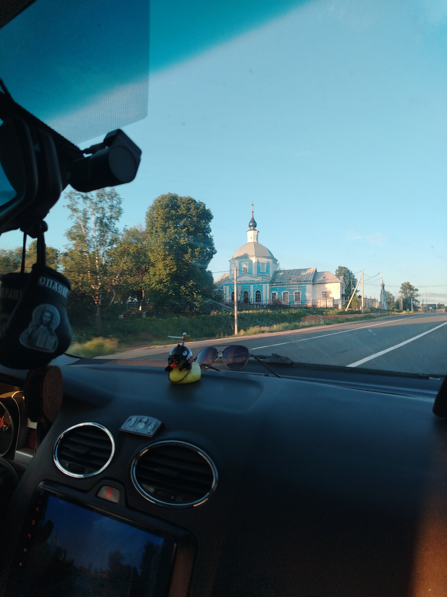 The image, taken from the interior of a car, shows a road leading towards a blue and white church in Глебовское, Russia. A rubber duck and sunglasses rest on the dashboard of the car. A small, dark-colored religious icon hangs from the rearview mirror. The sky is clear blue. The road has white lines marking the lanes. Trees line the left side of the road.