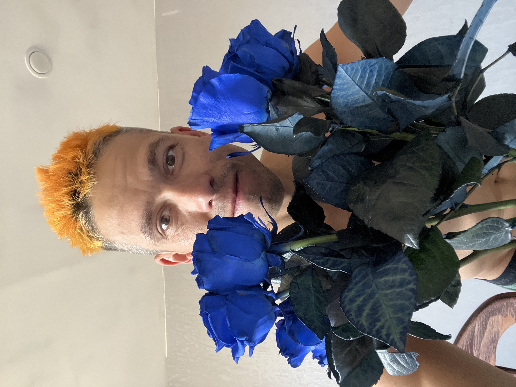 The image shows a person holding a bouquet of blue roses. The person has bright orange hair and is looking at the camera. The background is a plain wall. The image was taken indoors, likely in a residential setting. There is no visible indication of the city of Chaikovsky, Russia.