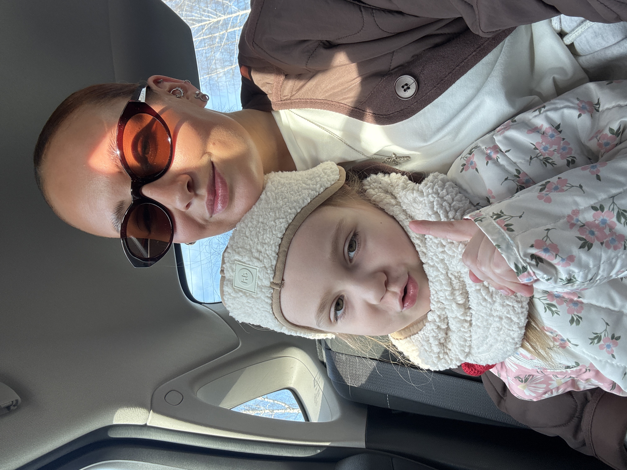 Inside a vehicle in an unspecified location of Chaikovsky, Russia, a woman wearing sunglasses and a brown jacket, sits with a young girl dressed in a floral jacket and a white fluffy hat. The interior of the car, including the headliner and window frame, is visible. Bare trees can be seen through the window. The girl is making a hand gesture.