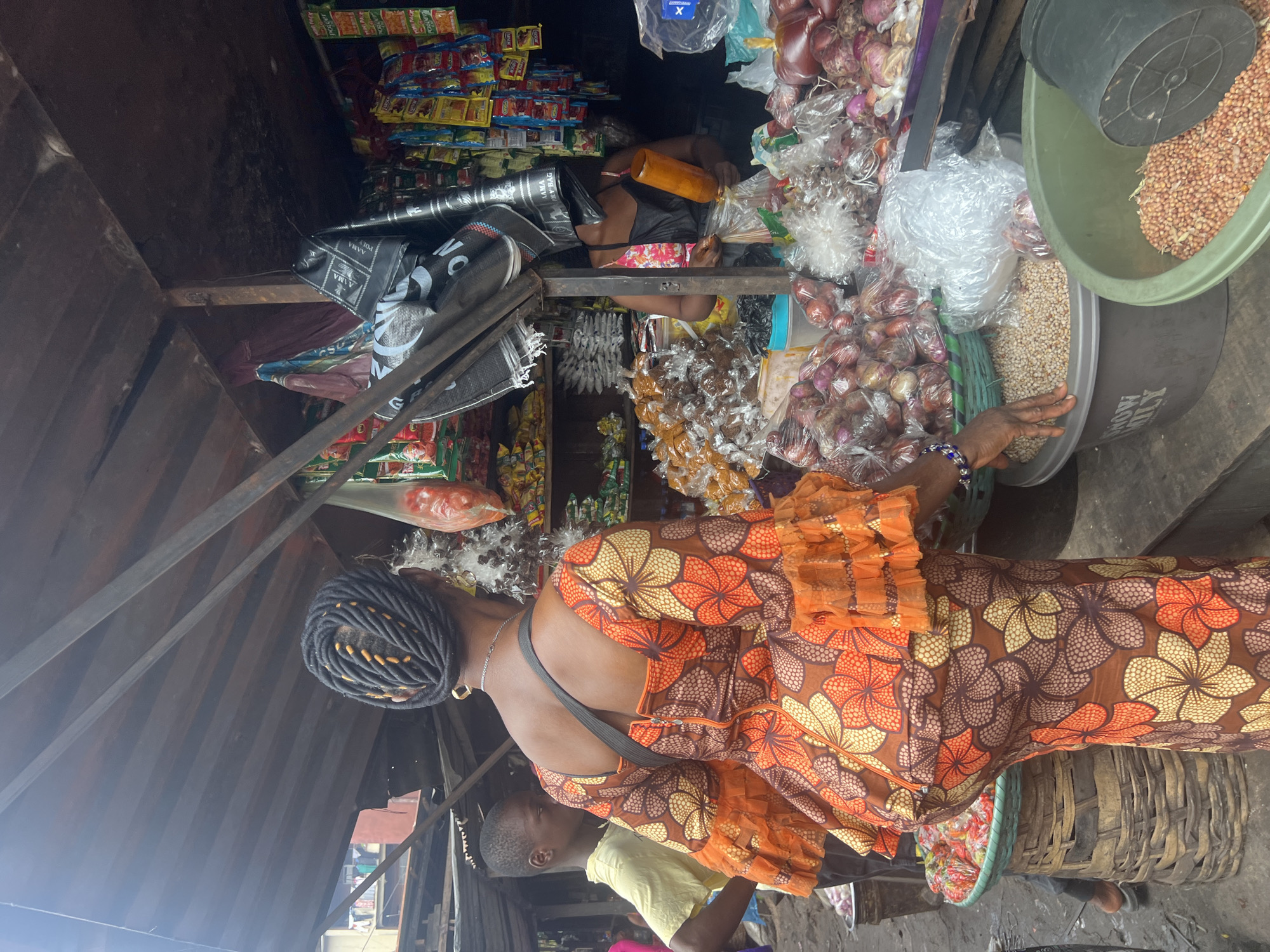 A woman wearing an orange, brown, and yellow floral print dress with ruffled sleeves is standing in a market stall in Nkpor, Nigeria. Her dark hair is styled in intricate braids with yellow accents. She is facing away from the camera, with her left arm slightly bent and her hand resting on a large grey tub filled with small, pale, round items. To her left, a young boy in a yellow t-shirt stands looking forward. The market stall is covered by a corrugated metal roof and filled with various goods, including bags of onions in plastic wrapping, baskets of produce, and shelves stocked with packaged snacks and condiments. A woman in the background, partially obscured by a black bag, is holding a cylindrical object and appears to be interacting with a customer. The ground is dirt and debris.