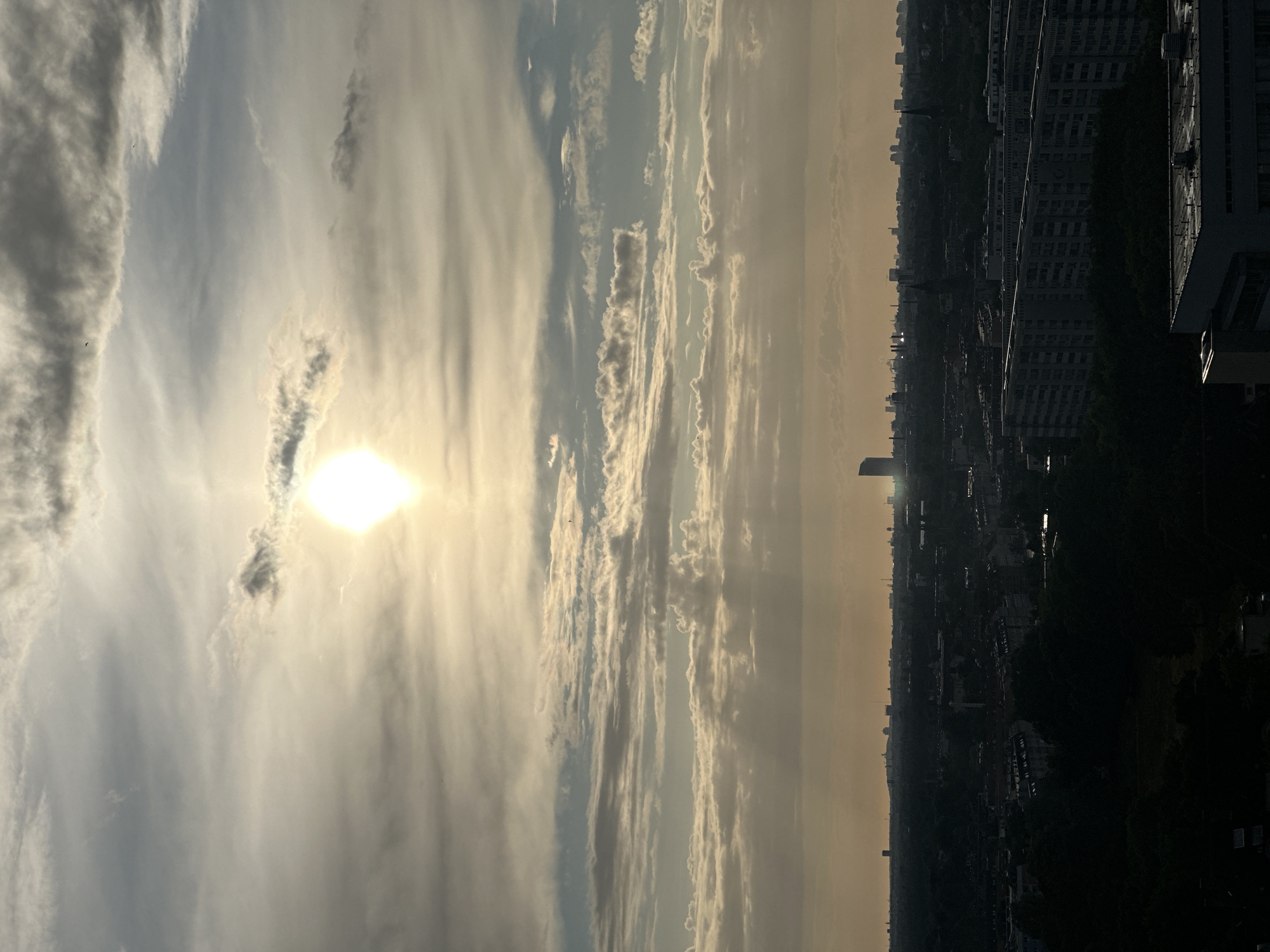 A panoramic view of a city skyline at sunset is depicted. The sun, a bright orb, is positioned slightly left of the center of the sky, casting a warm glow through scattered clouds. Rays of light descend through the atmosphere, illuminating parts of the cityscape below. The city stretches across the horizon, appearing dark and silhouetted against the lighter sky. Various buildings, including apartment blocks and a prominent skyscraper in the distance, are visible. The foreground is dominated by the dark outlines of trees and buildings, suggesting an elevated vantage point looking down onto the urban landscape. The overall impression is one of a tranquil, atmospheric end to the day in a densely populated urban area.