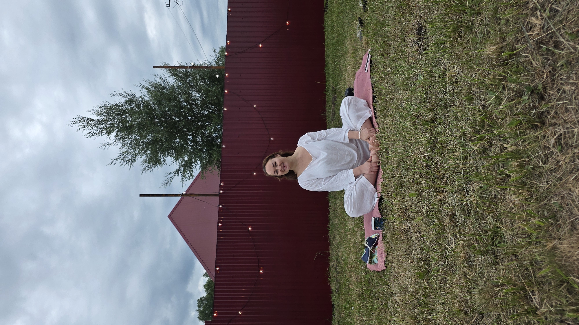 A woman is sitting in a meditative pose on a pink mat in a grassy yard. She is wearing a white outfit. A dark red corrugated metal fence is behind her, decorated with string lights. A red-roofed building is visible behind the fence. The sky is cloudy. The location is Kypoyarovo, Russia.