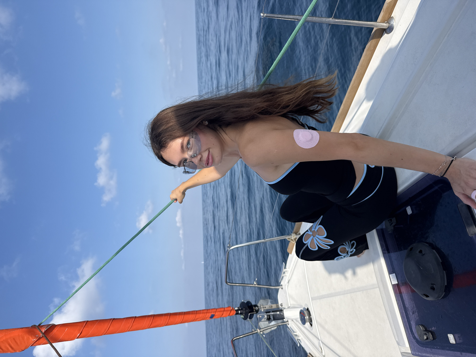 A young woman with long brown hair is sitting on the deck of a sailboat on a sunny day. She is wearing a black top with blue trim and black pants with blue floral designs. She has on blue-tinted sunglasses and a pink patch on her upper left arm. The sailboat is in the blue ocean, with a clear blue sky above, dotted with white clouds. The woman is holding onto a green rope connected to the boat's orange sail. The scene appears to be at sea, likely off the coast of Herzliya, Israel, given the context of the provided information, as the calm waters and clear sky are typical of the Mediterranean coast in this region.