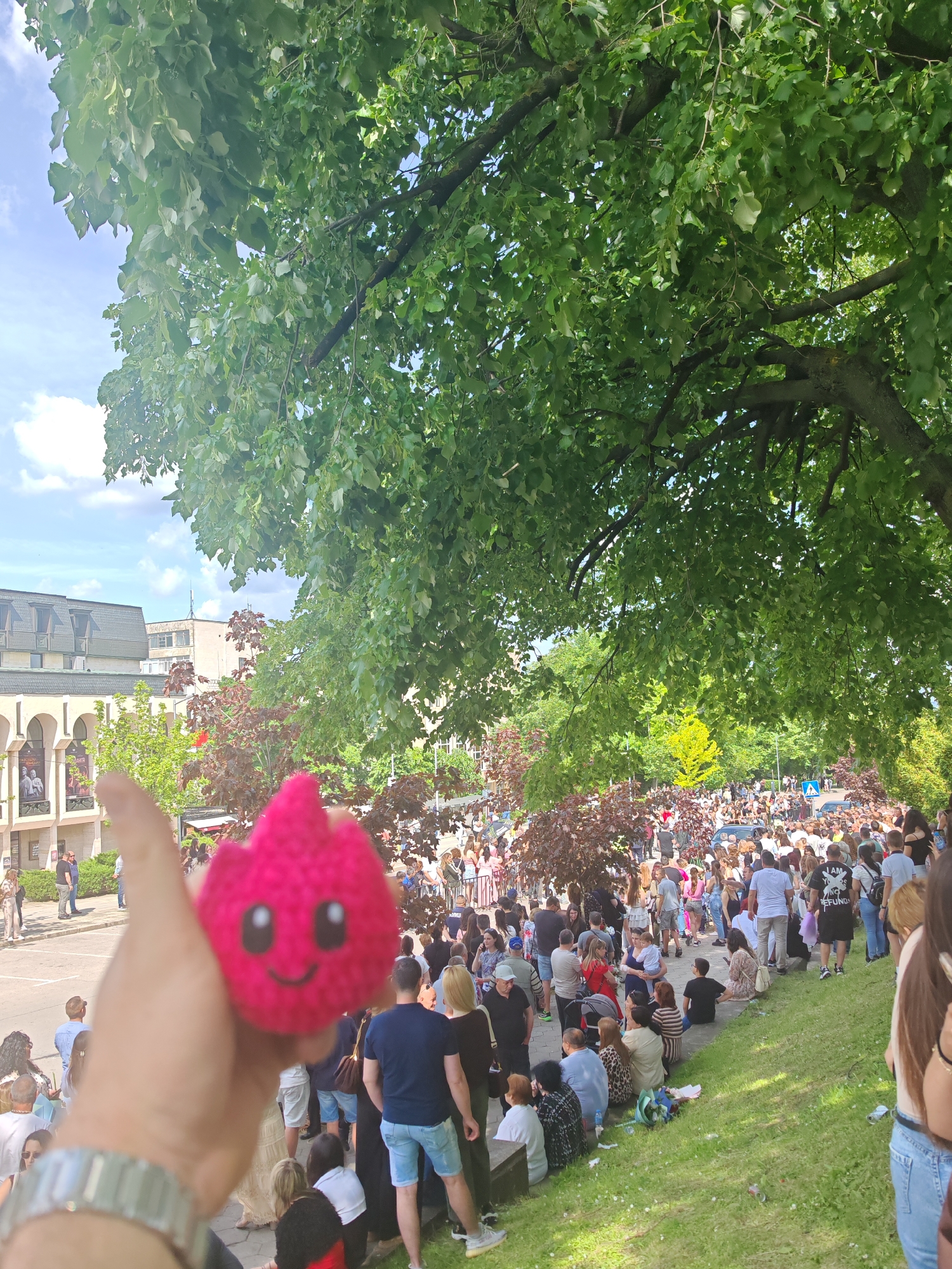 The image captures a scene outdoors in Shumen, Bulgaria. In the foreground, a hand holds up a small, pink, crocheted doll with a smiling face. Behind the hand and doll, a large crowd of people is visible, spread across a street and a grassy area. Many people are standing, while some are sitting on the grass. A tree with dense green foliage provides partial shade over the area. In the background, a building with classical architectural elements, including arches and a decorative facade, is visible. The scene suggests a gathering of people, possibly for an event, in an urban park-like setting.