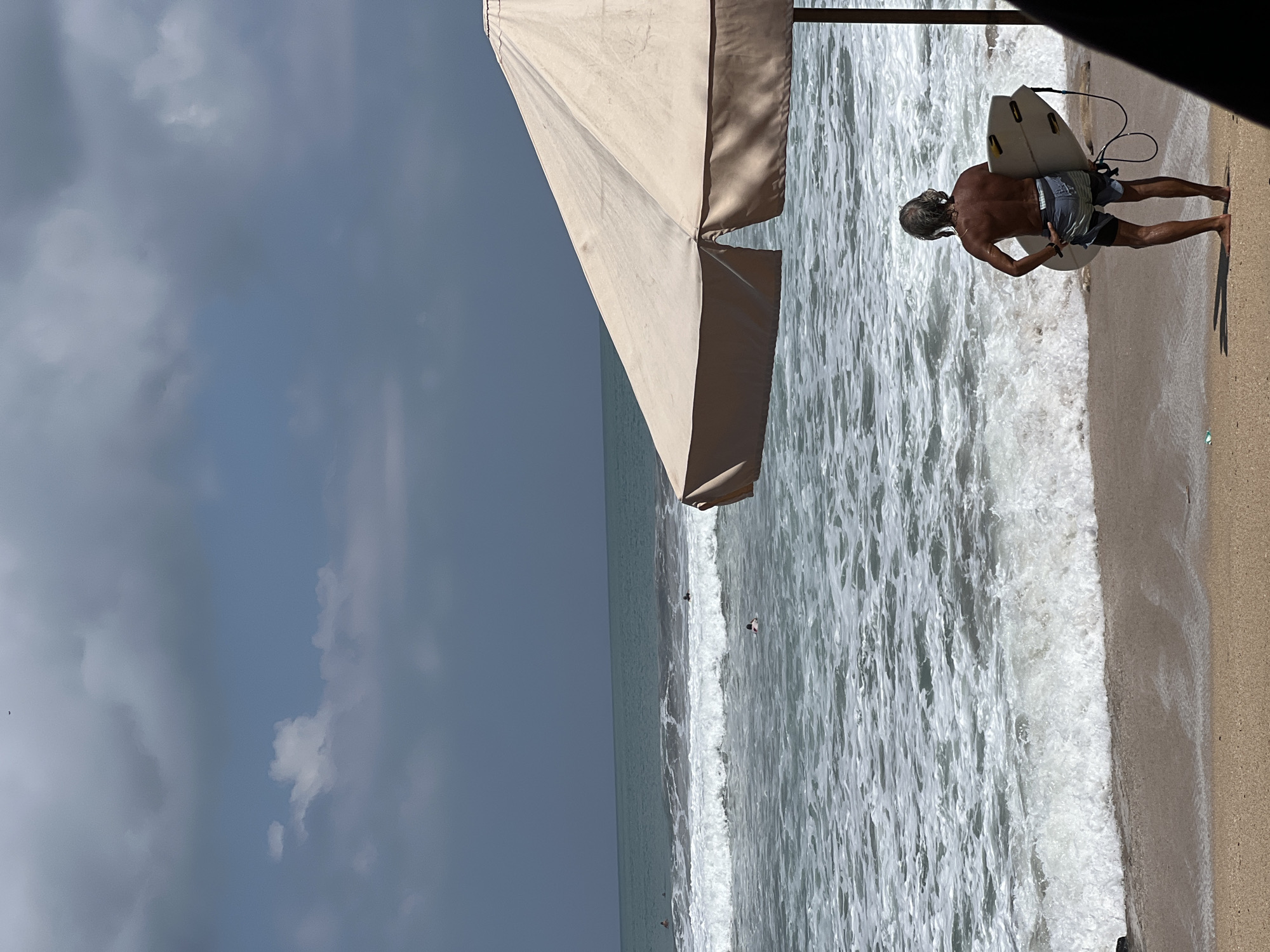 In Kuta, Indonesia, a man with gray hair stands on the sandy beach, facing the ocean. He is holding a white surfboard with his right hand on its side and his left hand at the bottom. The surfboard has a black leash attached. He is wearing blue and white striped shorts. The waves are crashing on the shore, and there are other surfers in the water. A large, beige beach umbrella is partially visible on the right side of the frame, providing shade. The sky is cloudy, with a mix of dark and light gray clouds. The ocean water is a greenish-blue color.