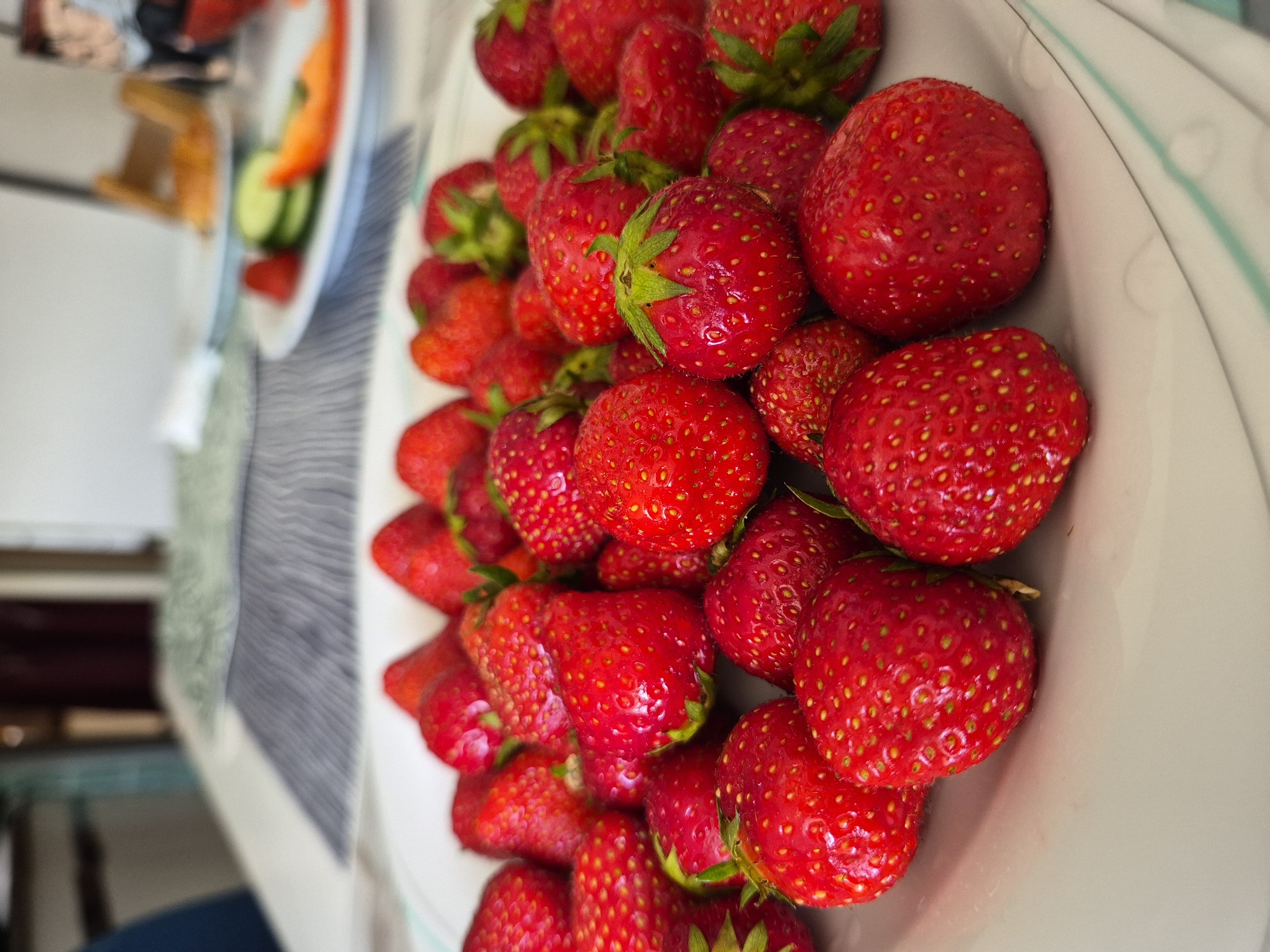 This is a close-up, overhead shot of a bowl filled with ripe strawberries. The strawberries are a vibrant red, with small yellow seeds visible on their surface. Some of the strawberries have green leaves attached to their stems. The bowl appears to be white with a light blue rim. In the blurred background, another plate with sliced vegetables, including cucumber and red pepper, is visible. A striped placemat is also partially in view. The lighting suggests it could be daytime, possibly indoors with natural light. The scene evokes a feeling of freshness and healthy eating. There is no visible text in the image.